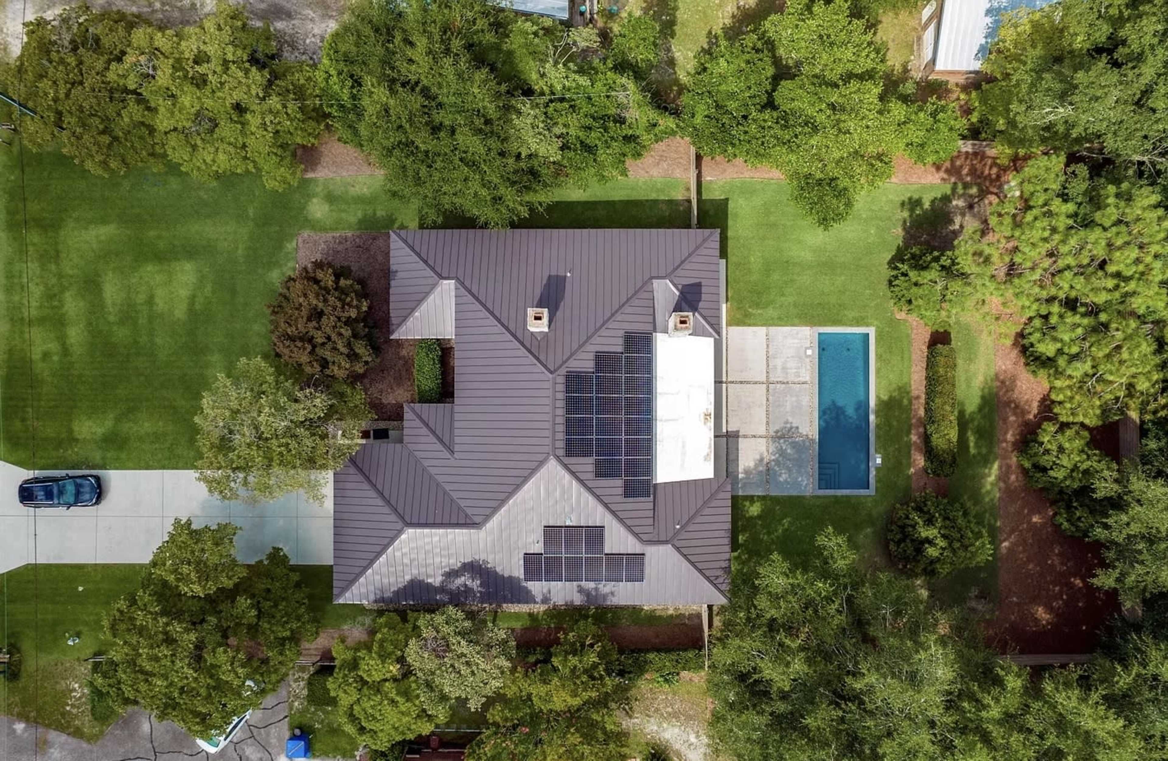 An aerial view of a house featuring a brown metal roof, solar panels, a pool, and surrounding lush greenery.