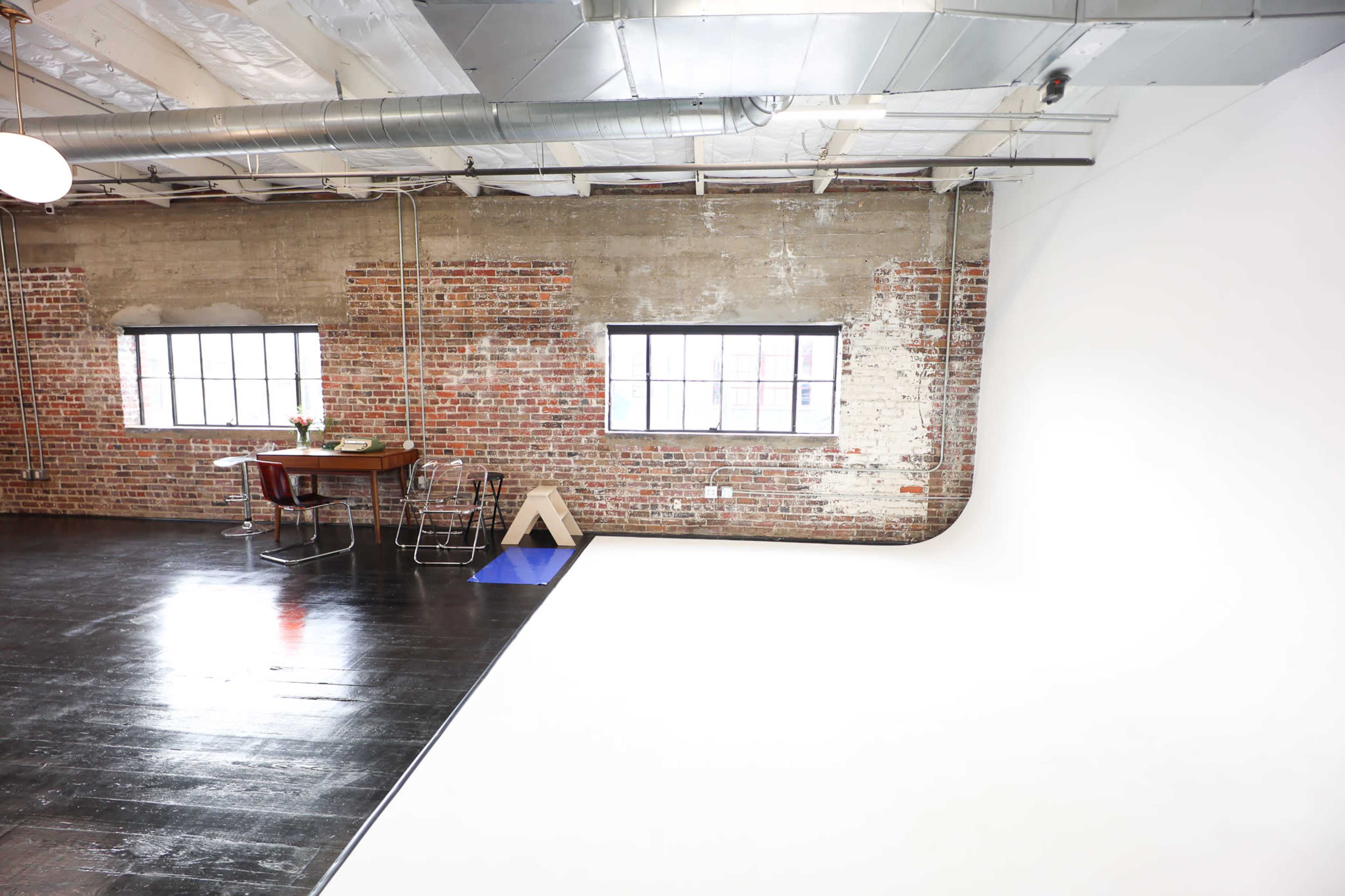 The image shows a spacious studio featuring exposed brick walls, large windows, and a clean white backdrop, with a dining table and chairs set off to the side.