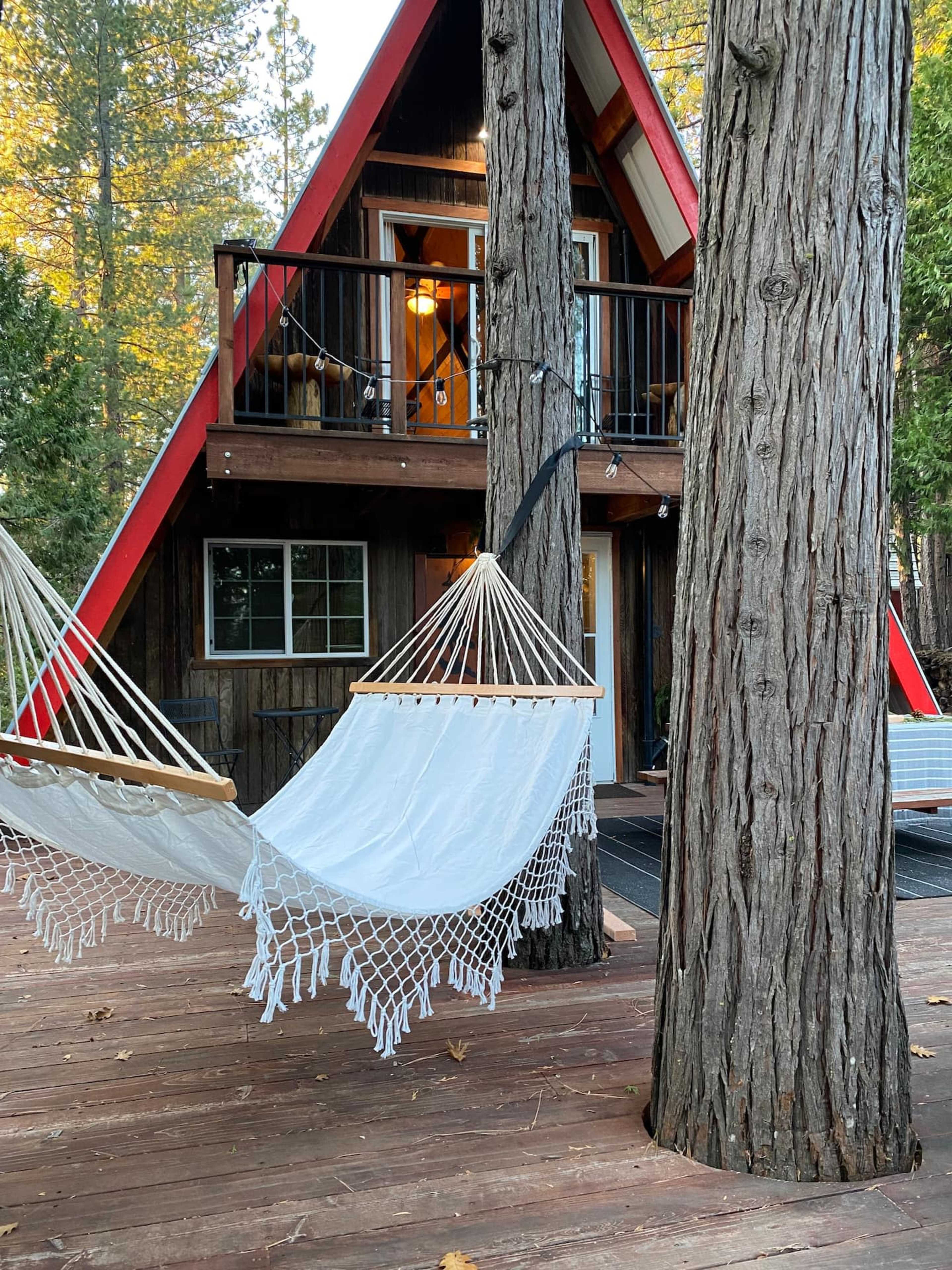A quaint A-frame cabin with red accents is situated among tall trees, featuring two hanging hammocks on a wooden deck.