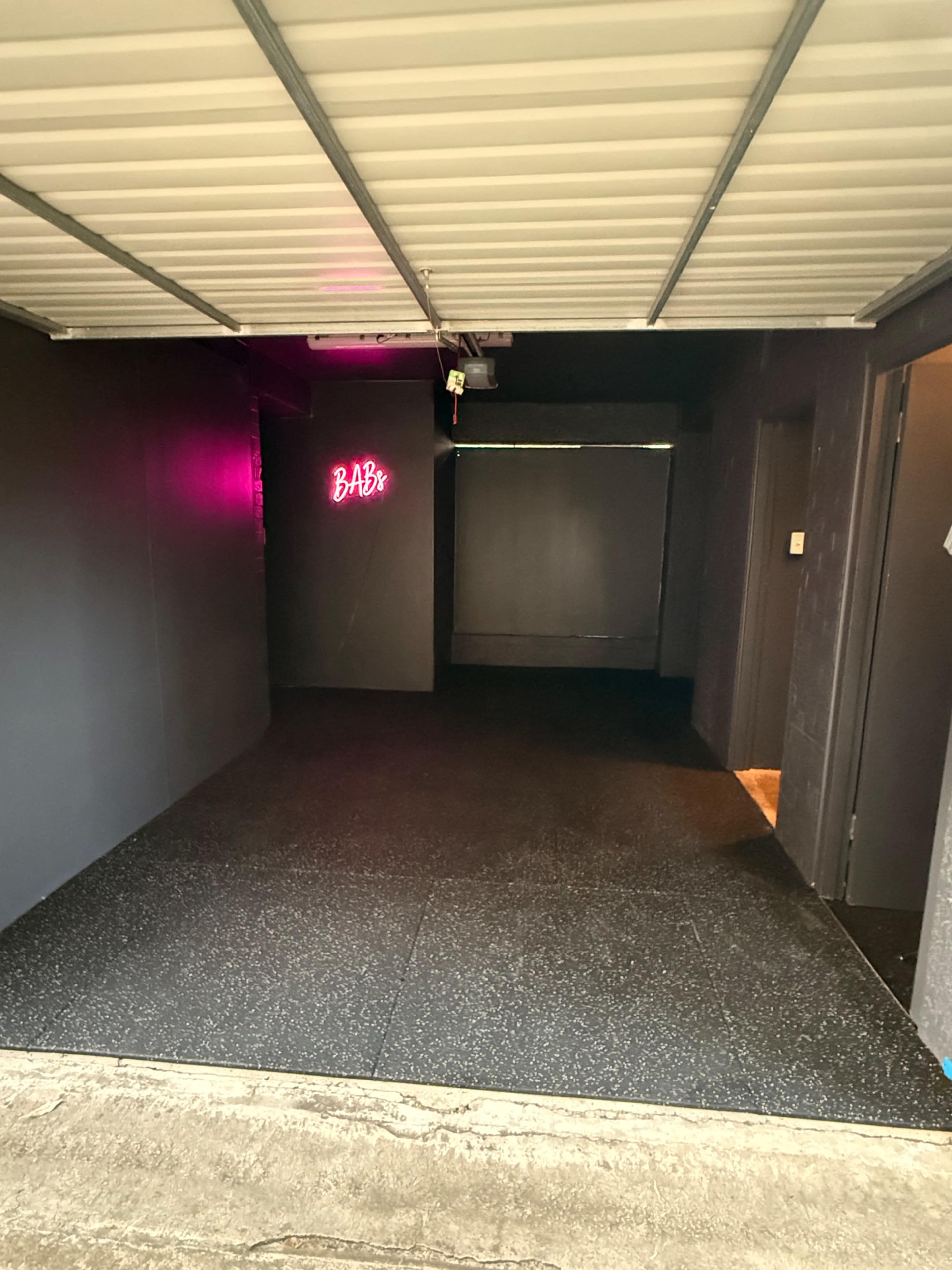 The image shows a dimly lit garage or studio space with black walls, a rubberized floor, and a neon sign that reads "BAB."