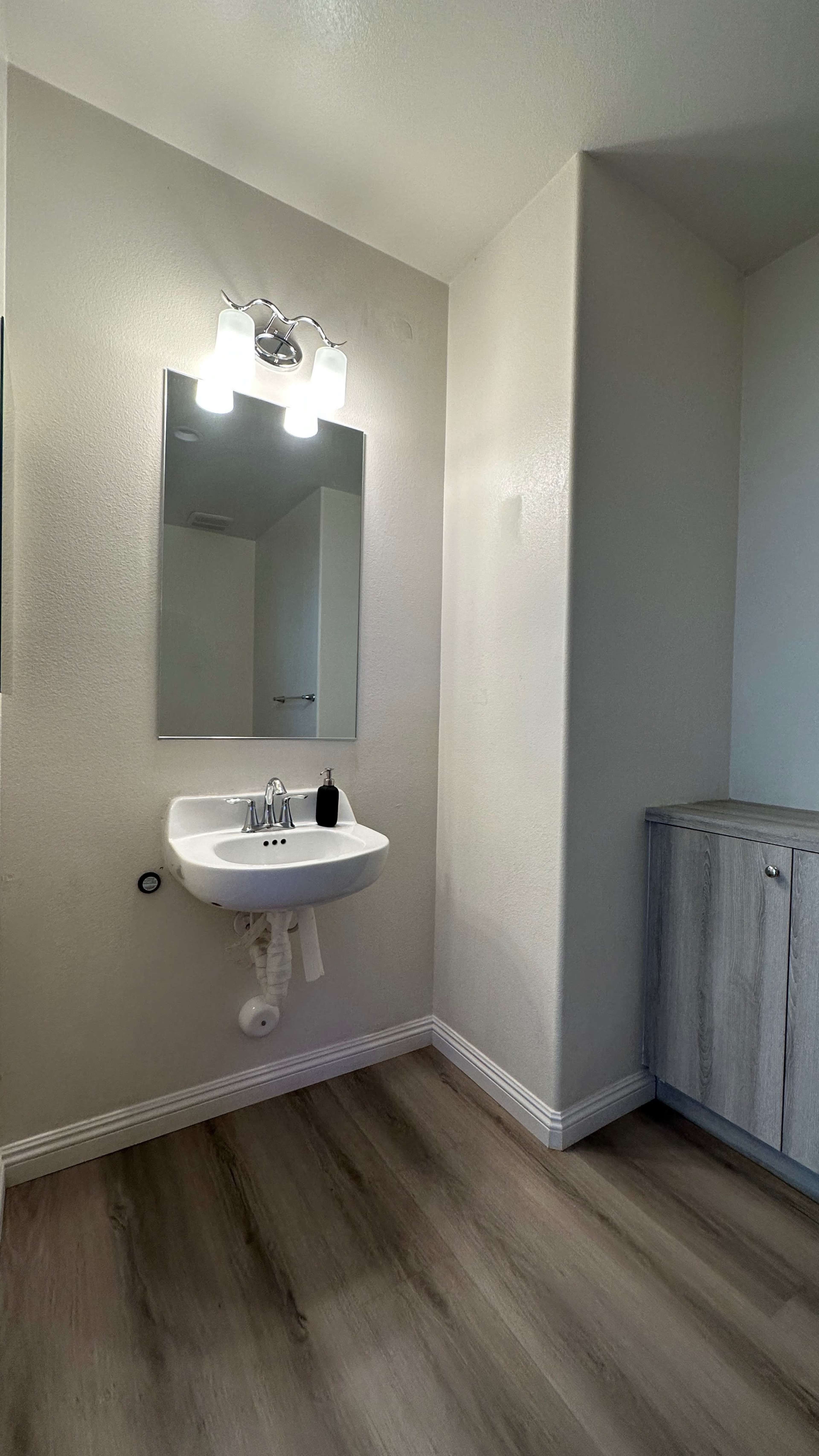 A small bathroom featuring a wall-mounted sink, a mirror, a light fixture above the sink, and a wooden cabinet in the corner.