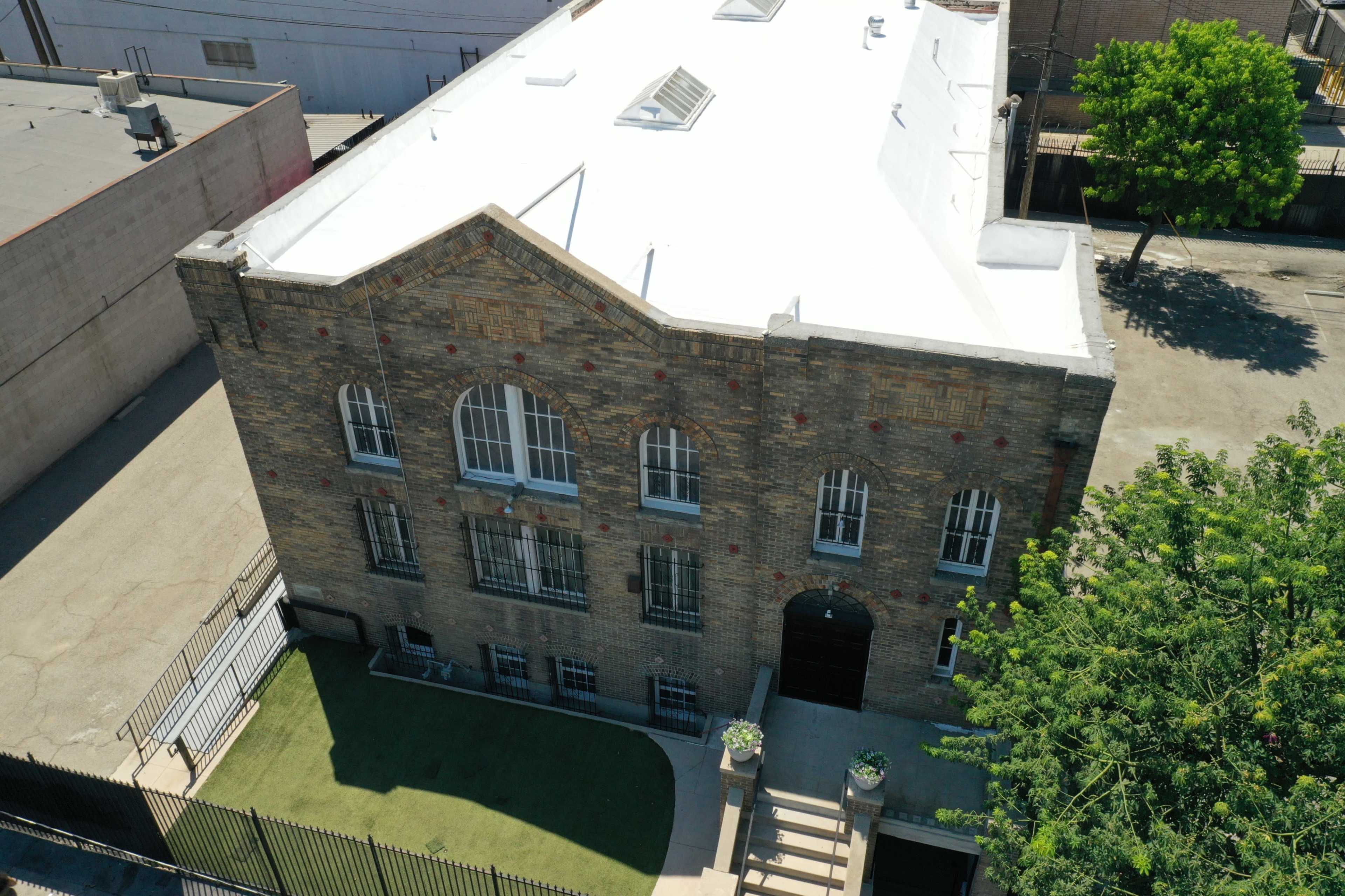The image shows a large, brick building with arched windows and a flat roof, situated next to a grassy area and bordered by a black fence.