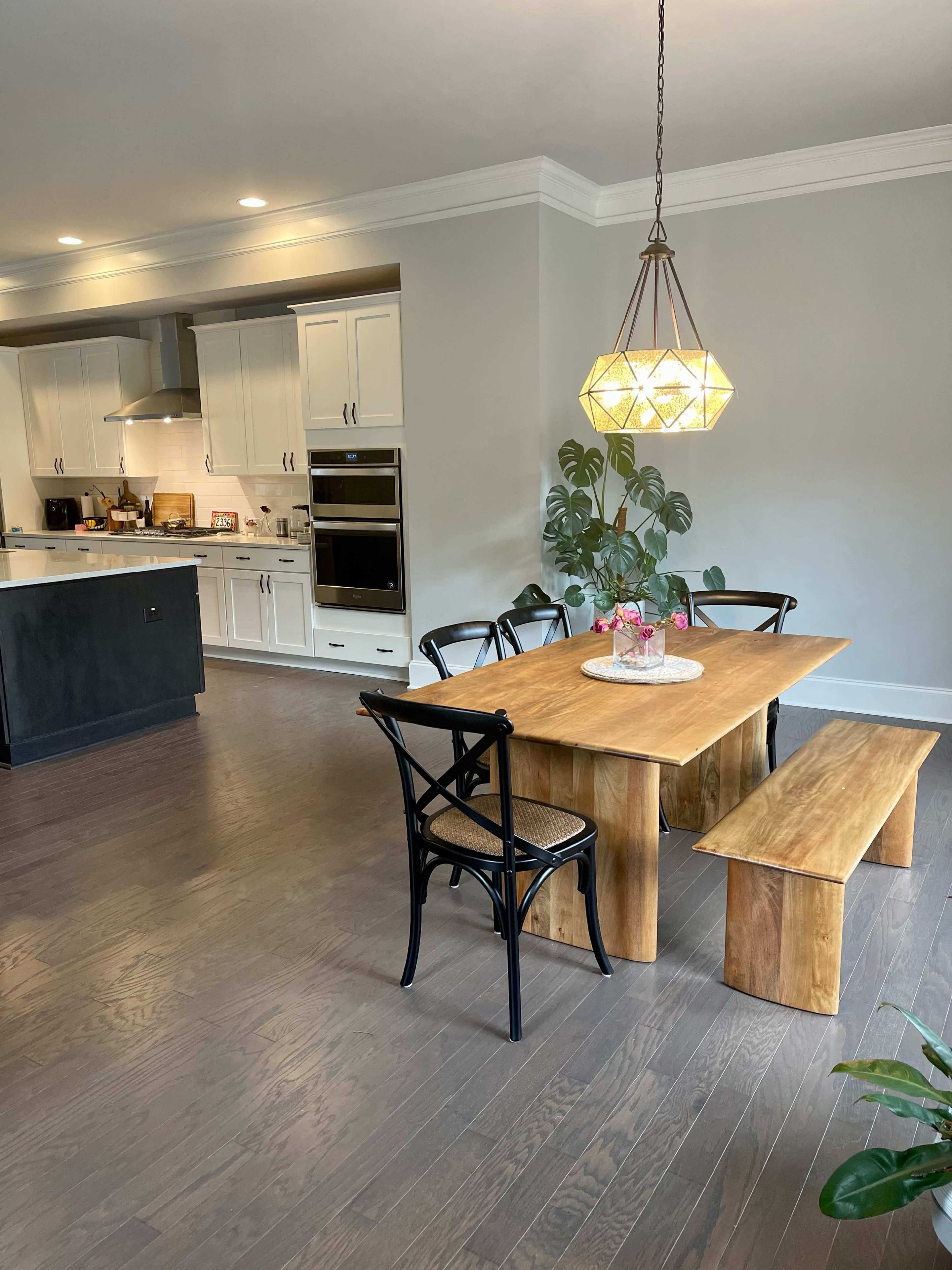A modern dining area with a wooden table, a bench, and a pendant light, adjacent to a kitchen featuring white cabinetry and a black island.