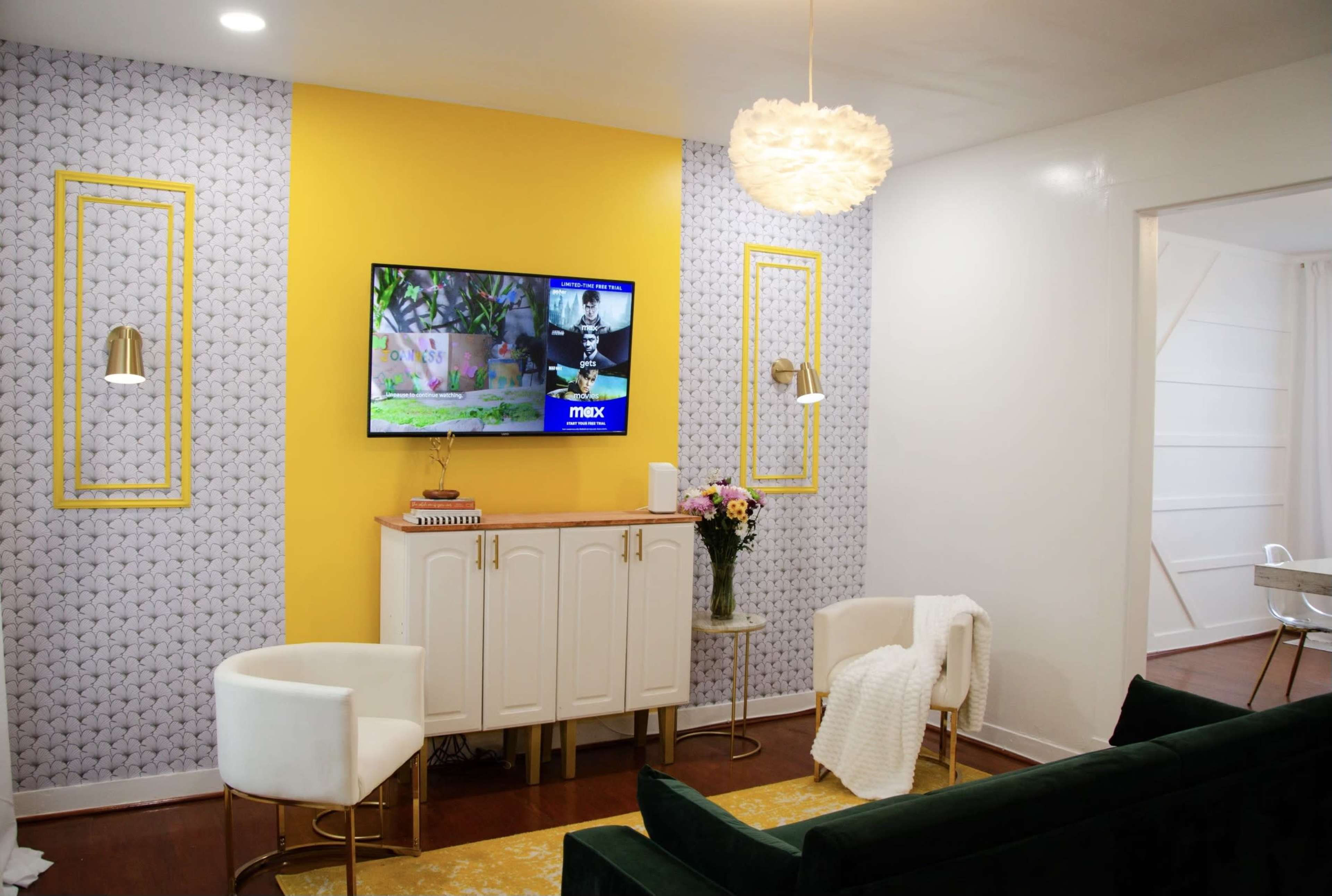 A modern living room with a yellow accent wall, a television mounted above a white sideboard, and two white chairs facing a green velvet sofa, complemented by floral decor.