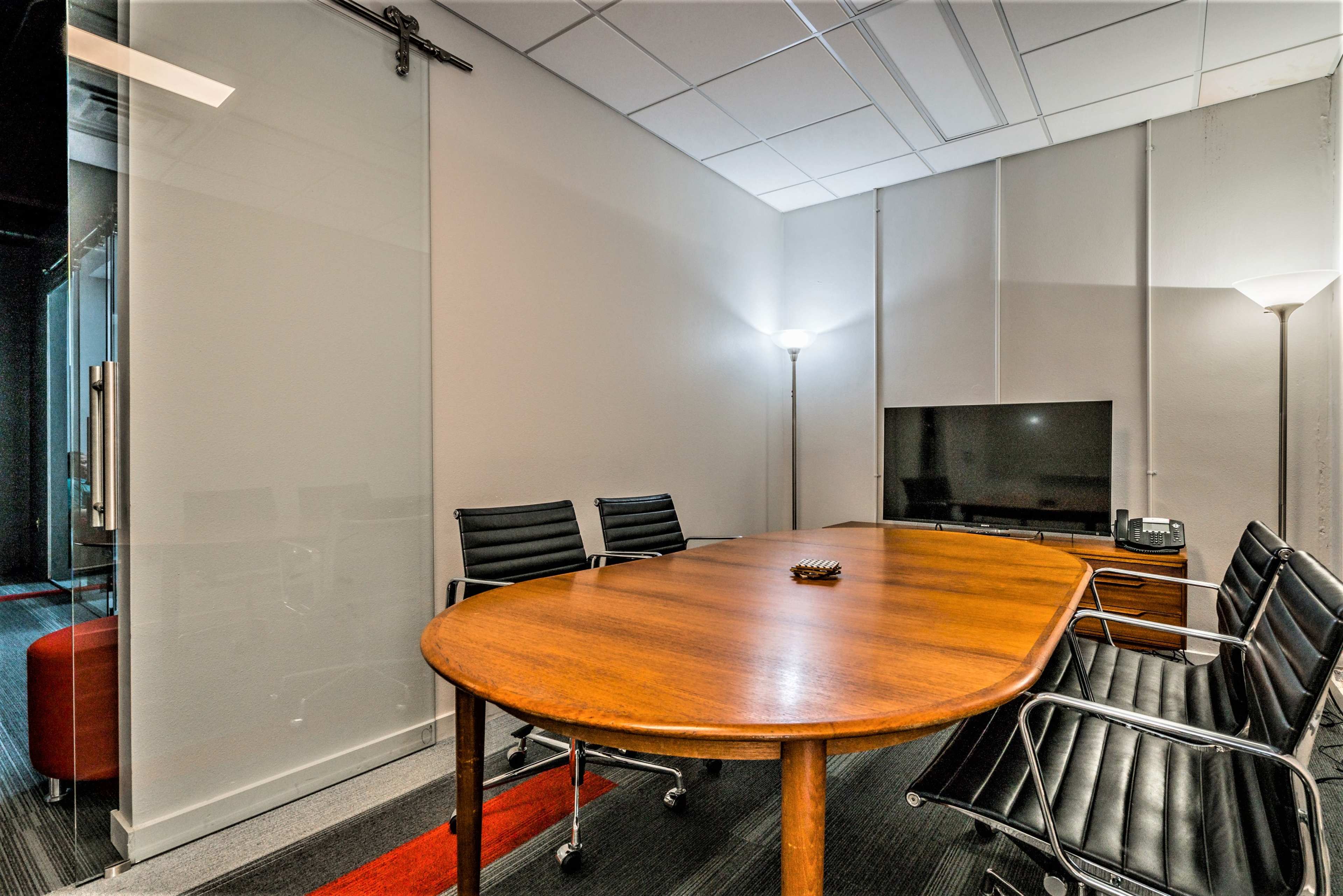 The image shows a modern conference room with a wooden oval table surrounded by black leather chairs, a television, and two floor lamps.