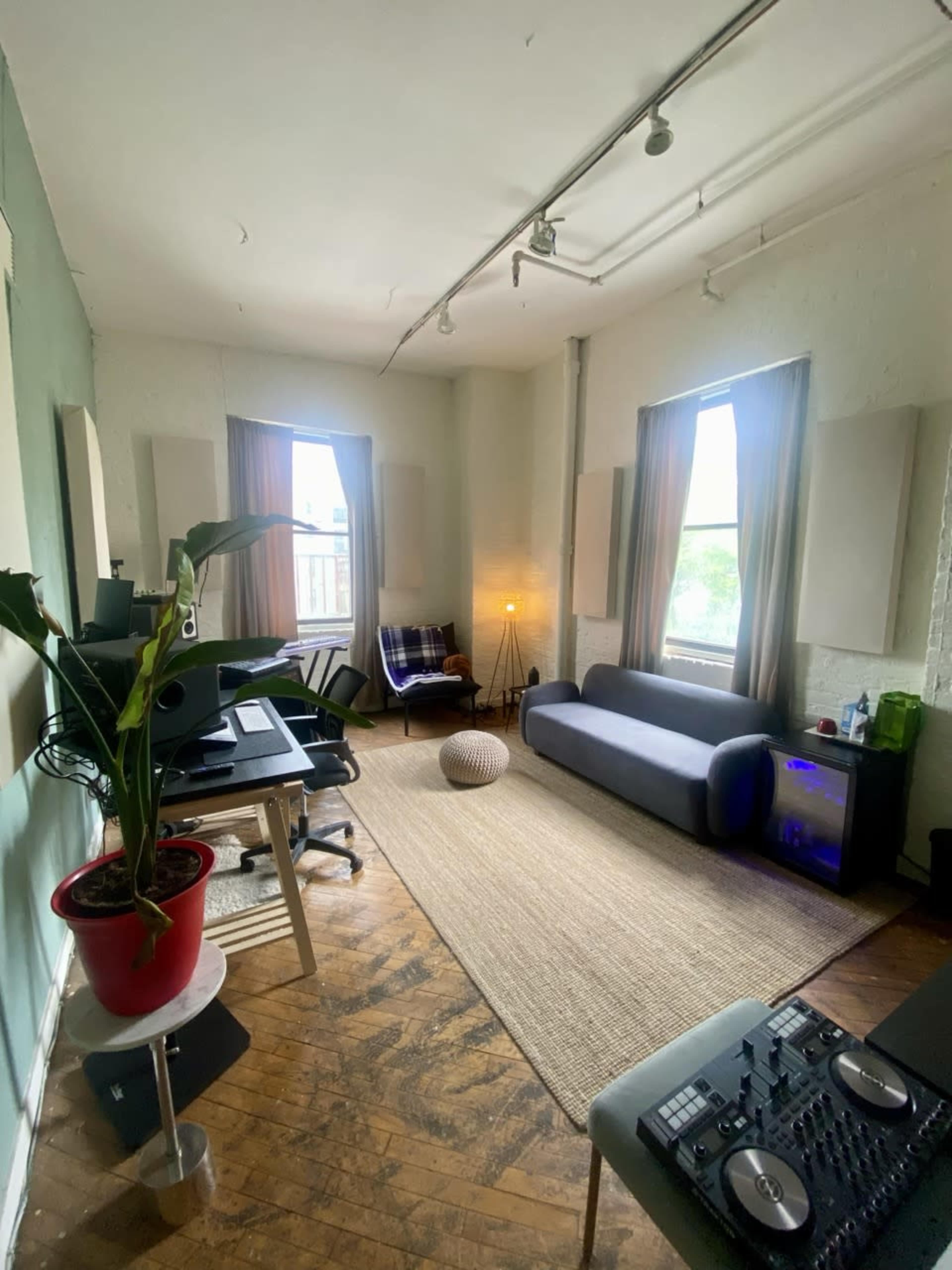 The image shows a spacious room with a sofa, a rug, a plant, workstations, and large windows providing natural light.