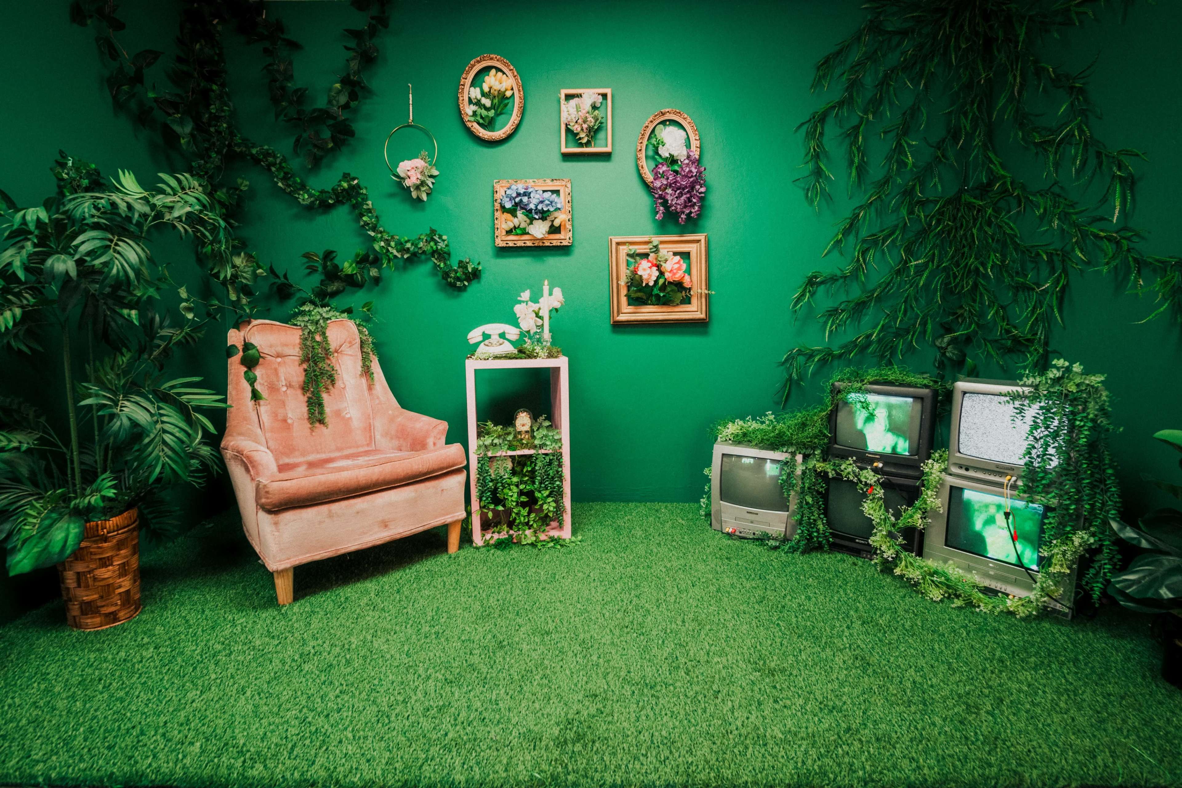 A pink upholstered chair sits against a vibrant green wall adorned with framed artwork and vintage televisions surrounded by decorative plants.