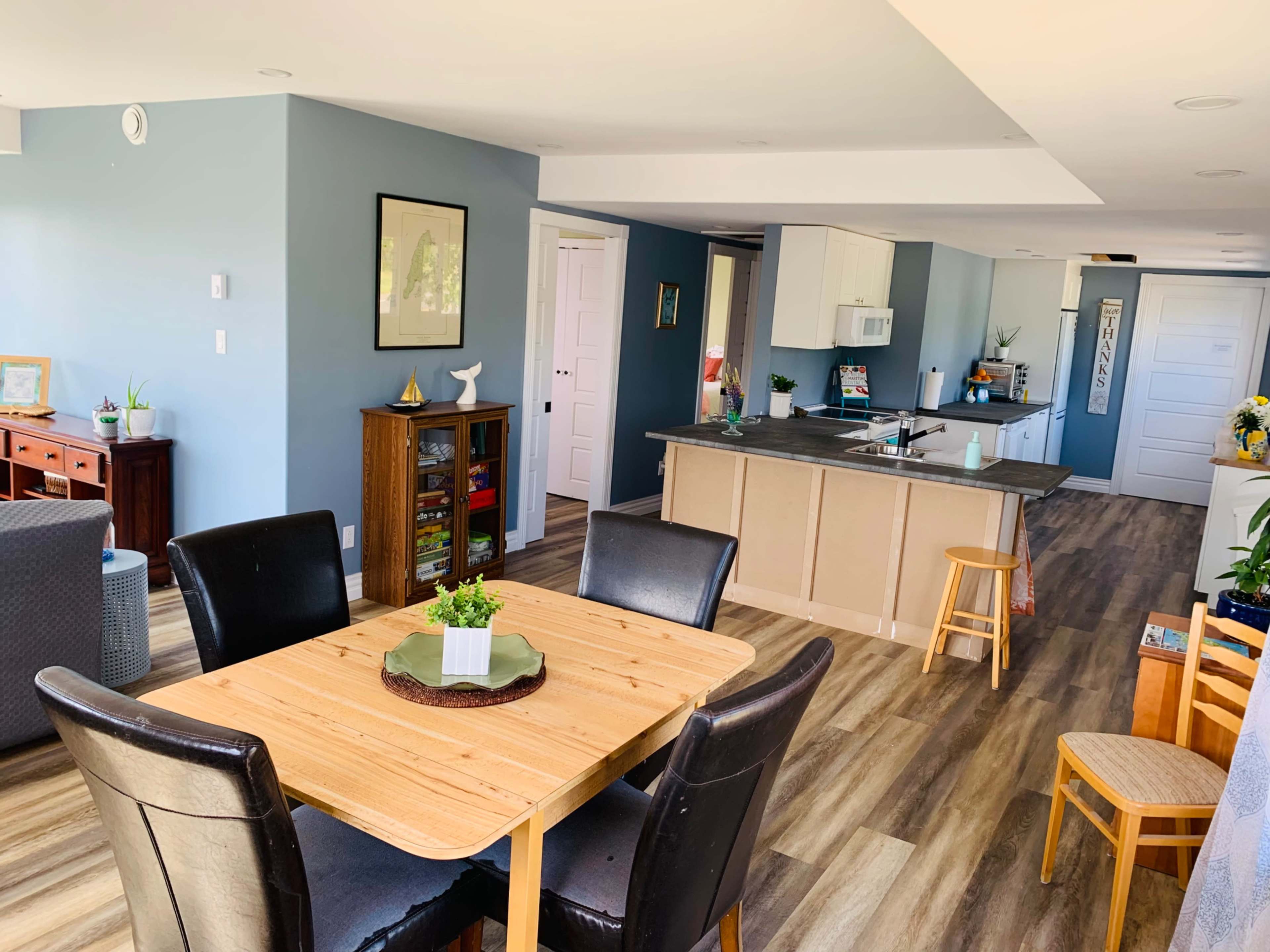 A spacious, open-concept living area featuring a wooden dining table surrounded by black chairs, a kitchen visible in the background, and light blue walls.