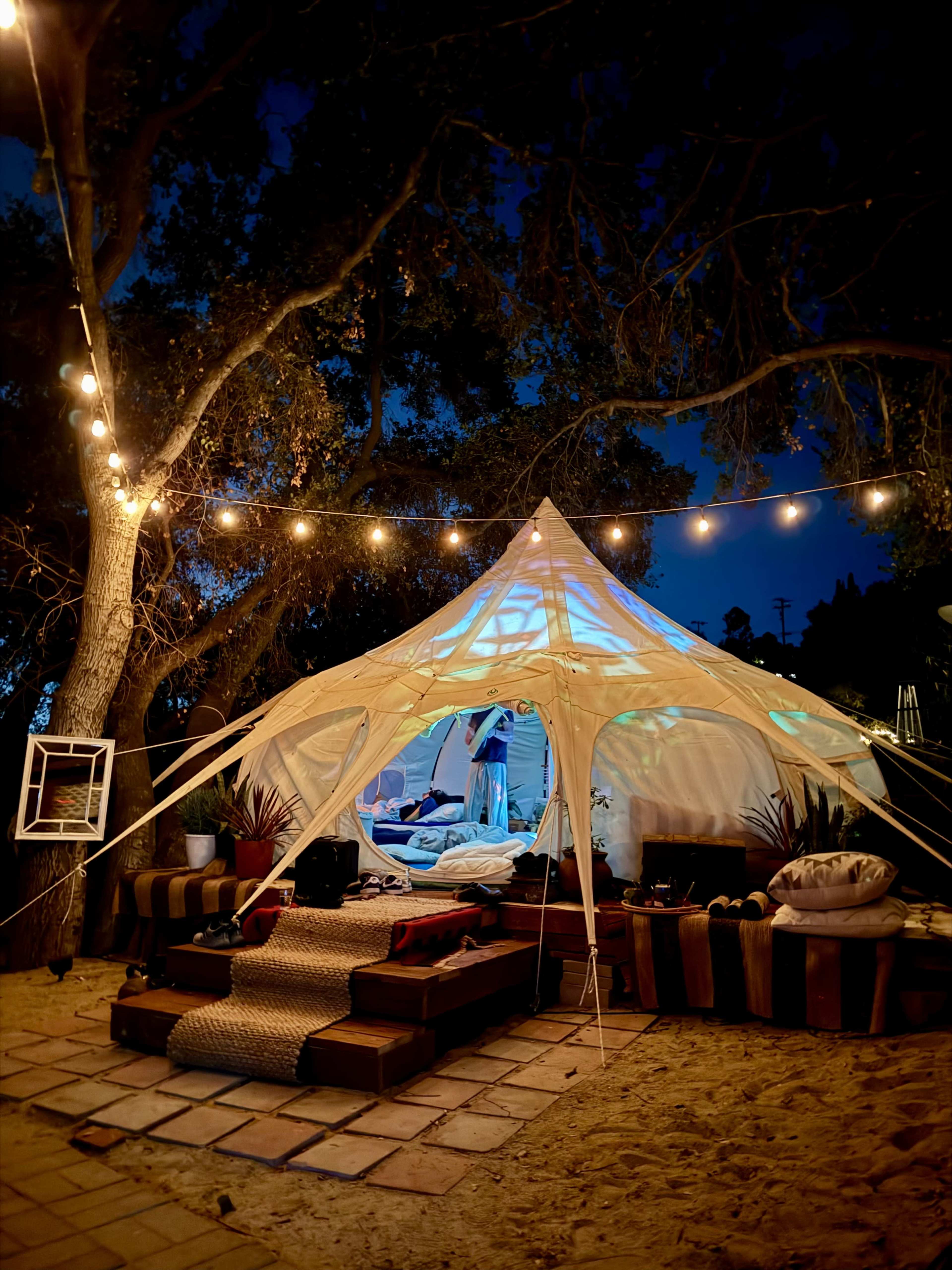 A large, illuminated tent is set up in a sandy area, surrounded by trees and string lights, with cozy seating and decorative elements.