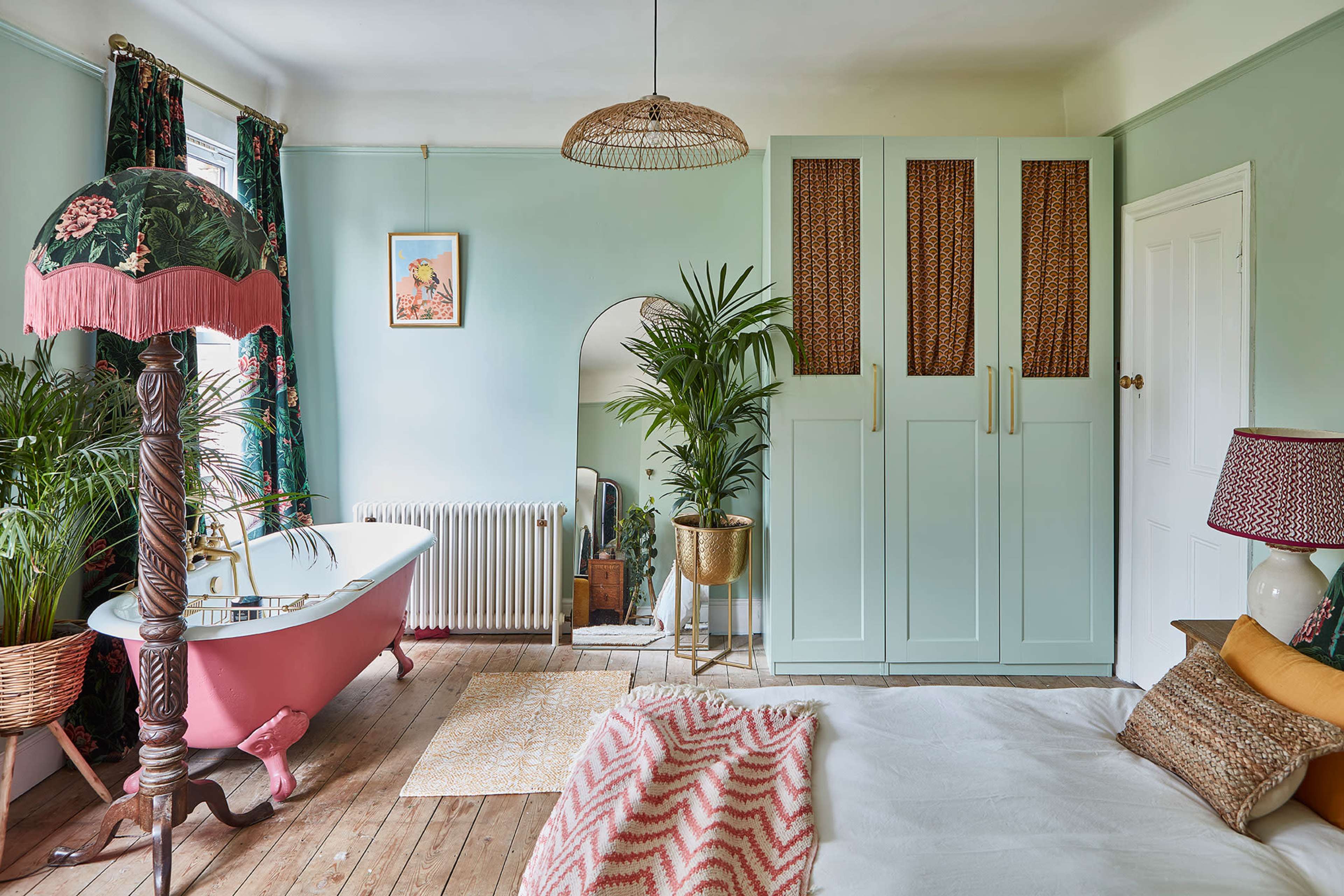 A softly lit bedroom featuring a pink bathtub, green walls, a large wardrobe, and a bed adorned with a patterned throw, accompanied by potted plants and decorative lamps.
