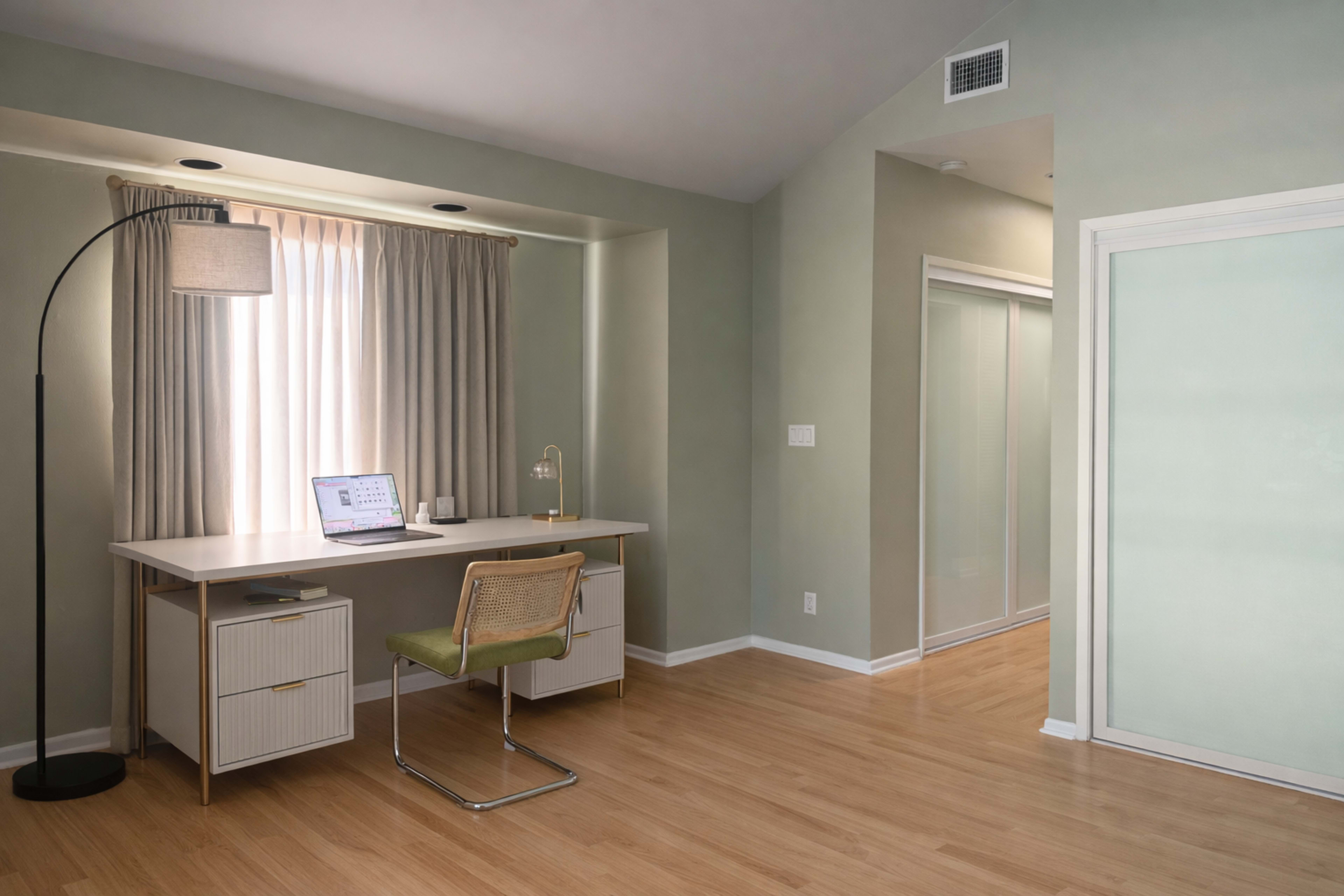 The image shows a modern, minimalistic home office with a desk, a chair, and a floor lamp, set against light green walls and wooden flooring.