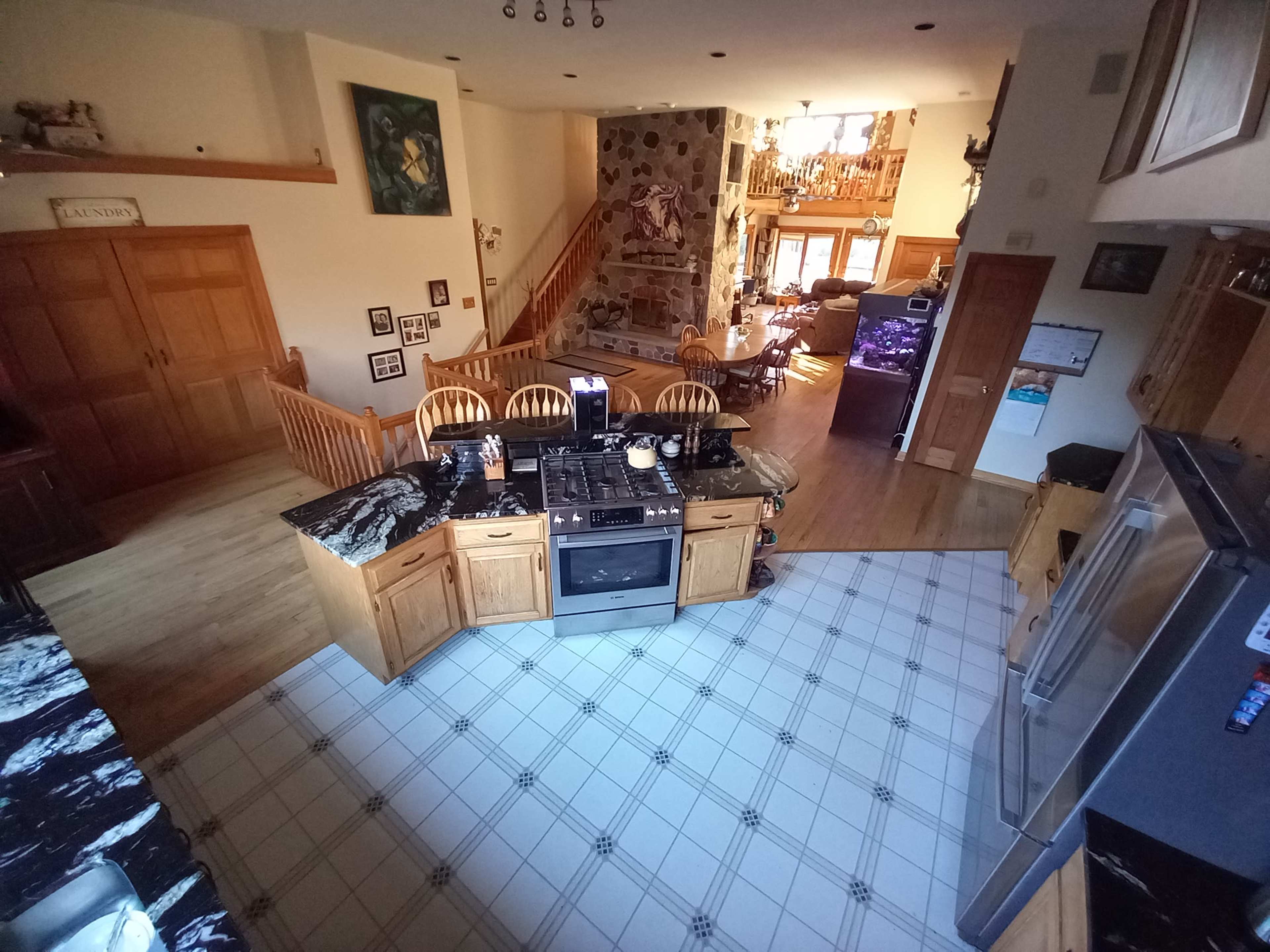 A spacious kitchen with a black and white tiled floor, wooden cabinets, and a central island, overlooking a bright living area with a staircase and a stone wall.