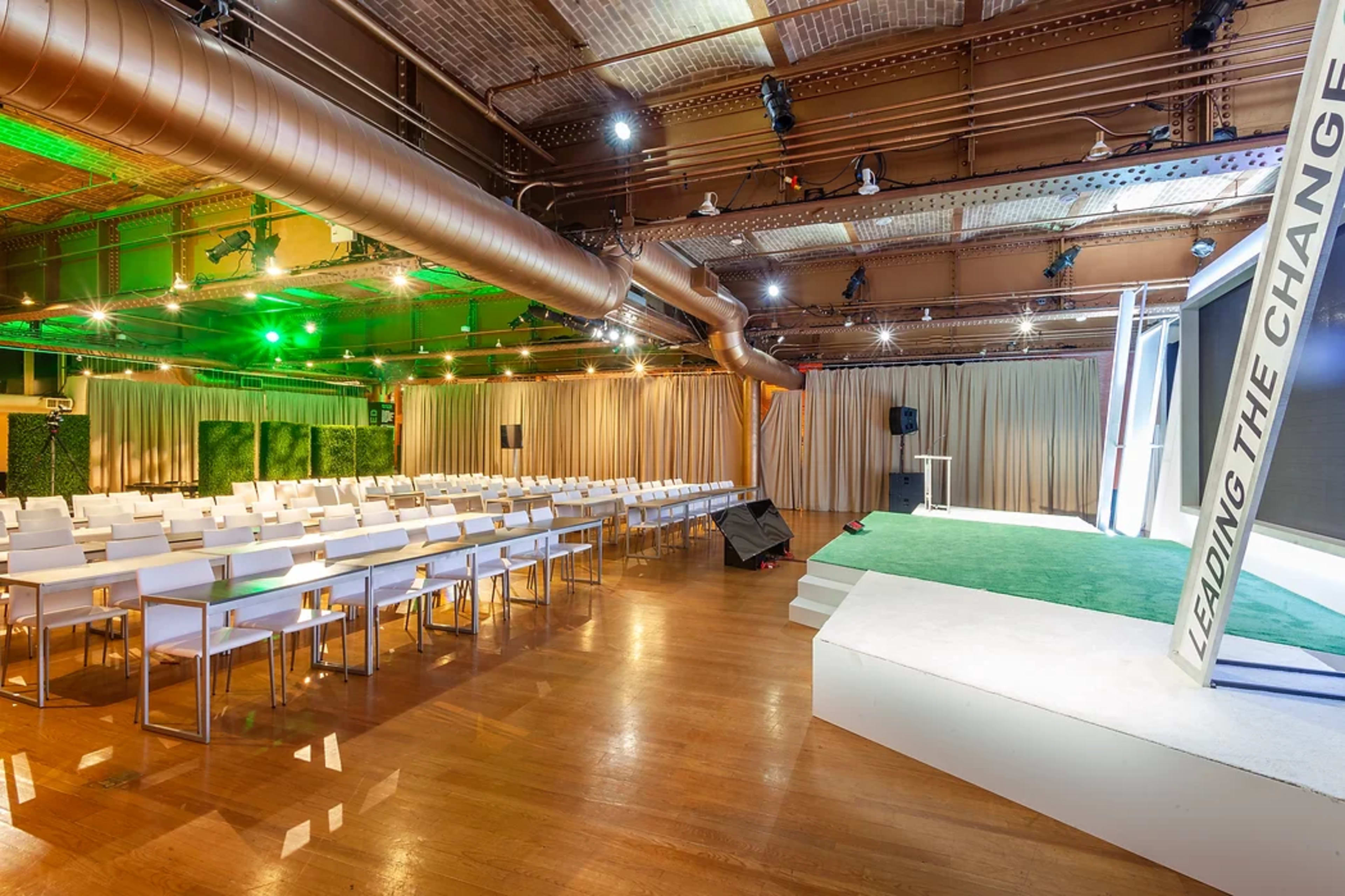 A spacious event venue features rows of white chairs facing a stage with a green carpet and large screens, surrounded by beige curtains and industrial-style decor.