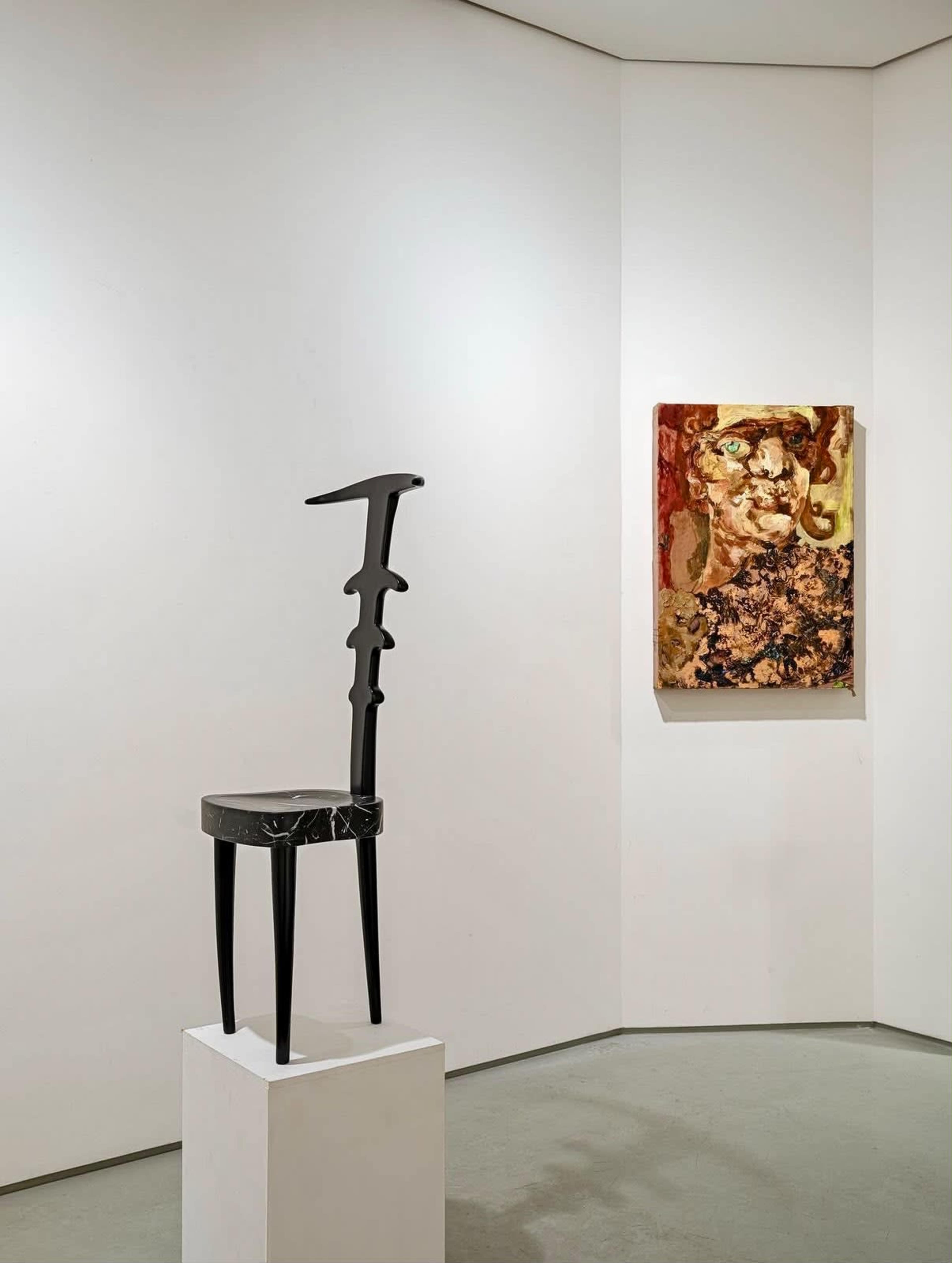A black sculptural chair stands on a pedestal in a corner gallery, next to an abstract painting with earthy tones.