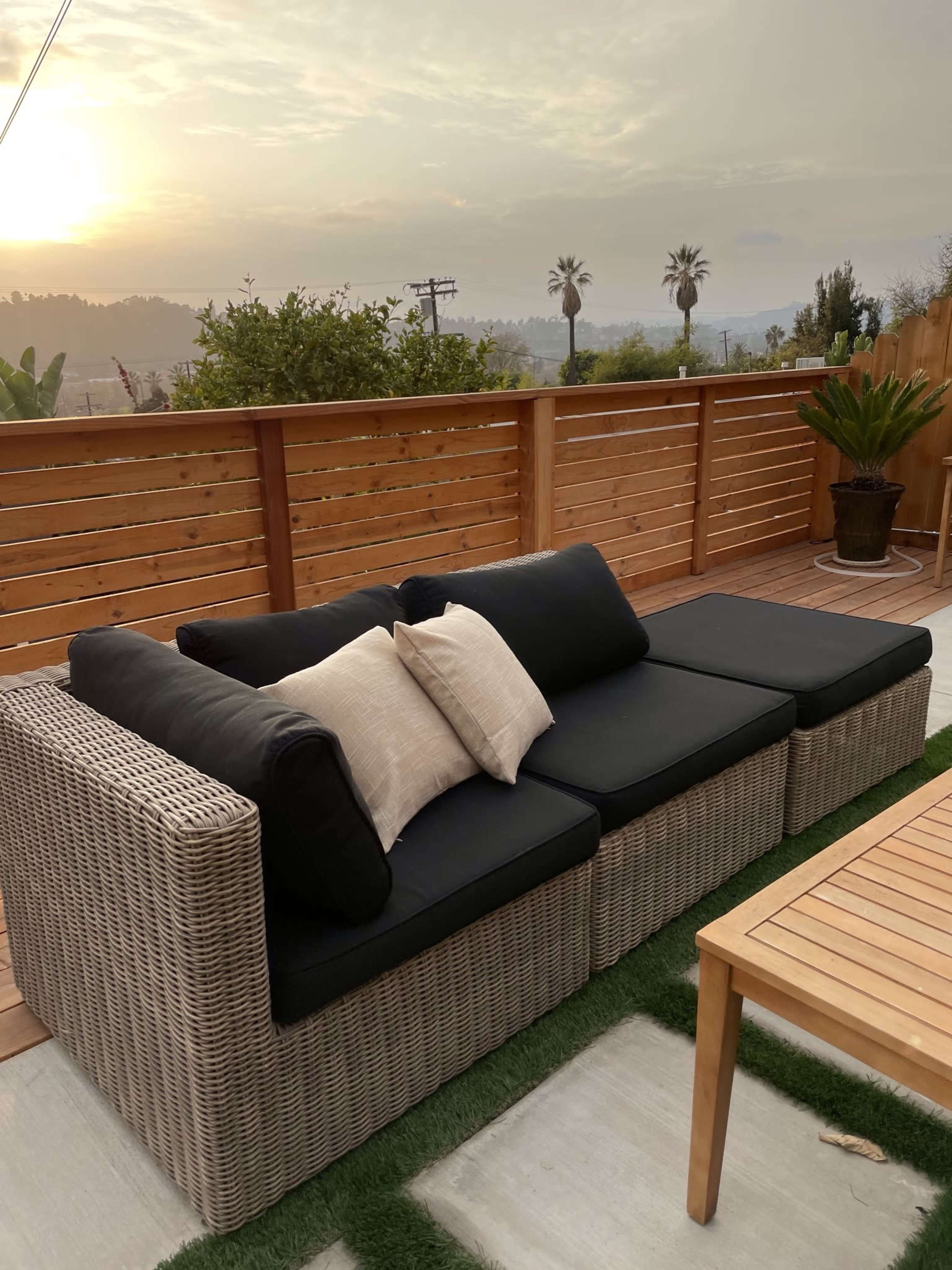 A wicker sofa with black cushions and a decorative pillow sits on a patio with a wooden fence and a view of palm trees against a sunset backdrop.