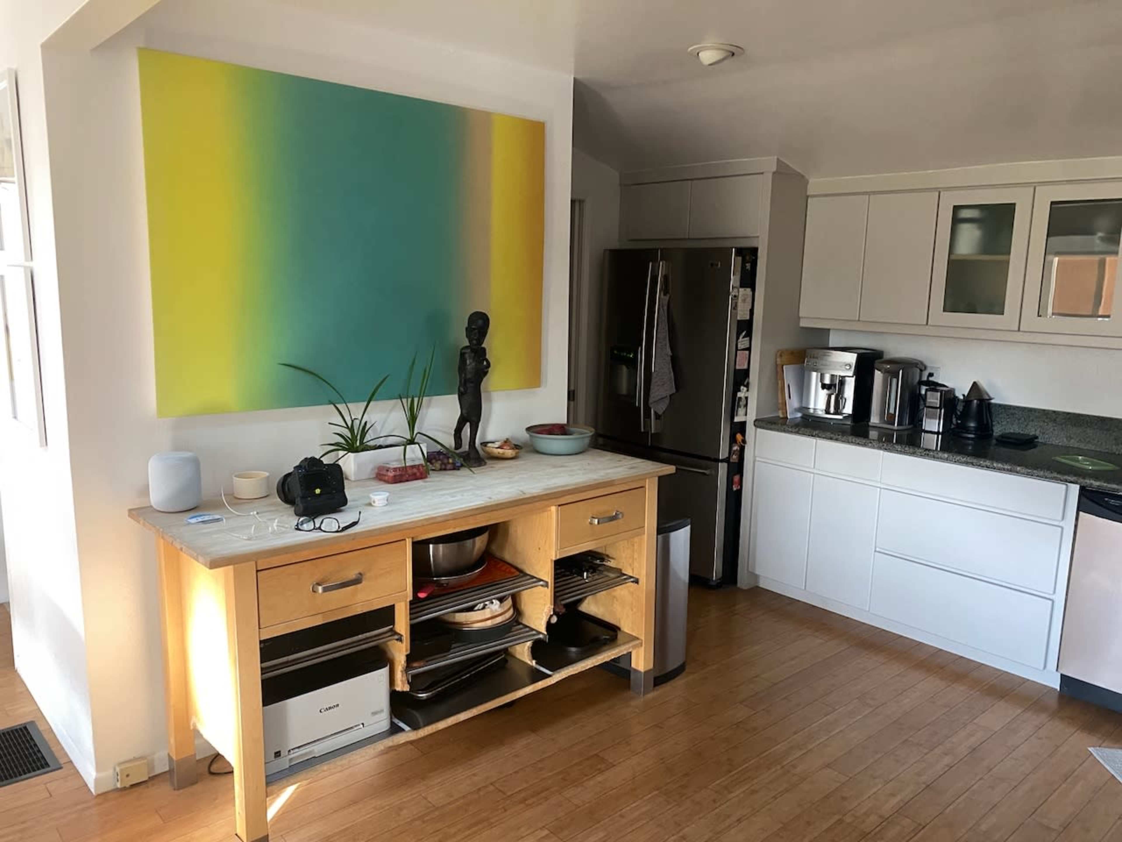 The image shows a modern kitchen with a marble-topped island, a refrigerator, and various kitchen appliances, along with a colorful painting on the wall and a small statue on the counter.