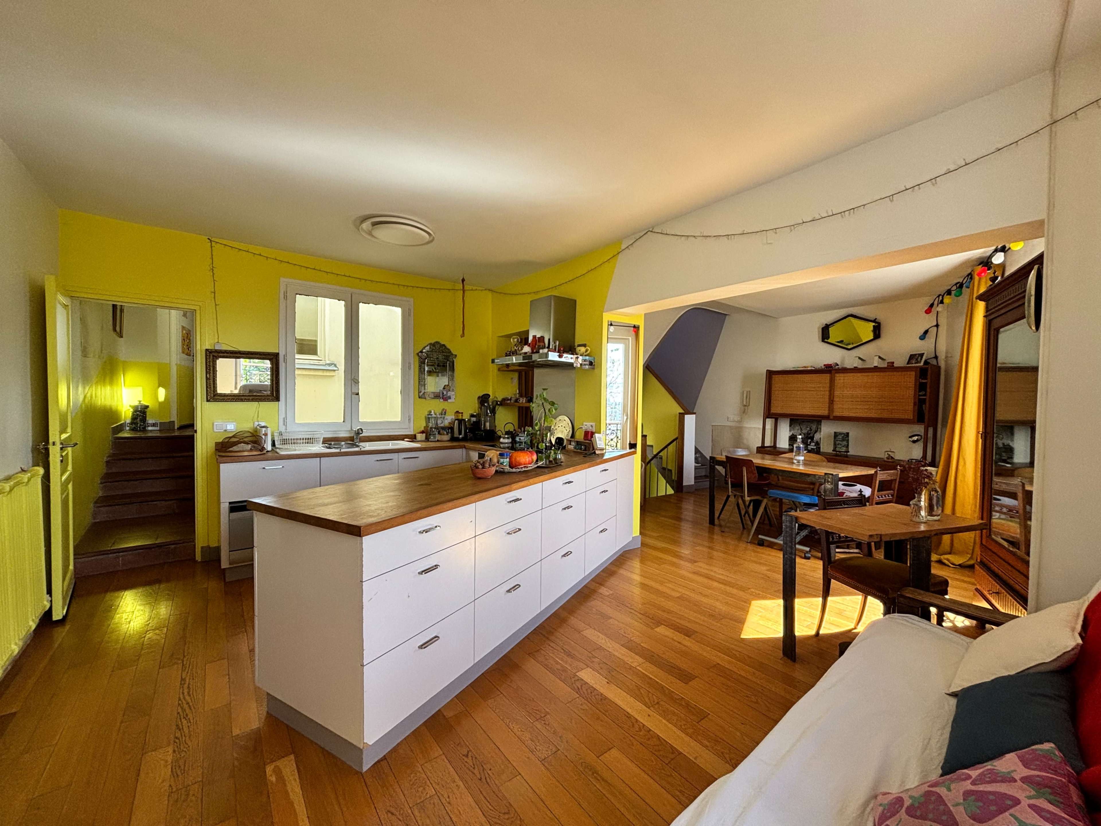 The image depicts a brightly lit kitchen and living area with wooden floors, a central island countertop, and a cozy dining space in the background.