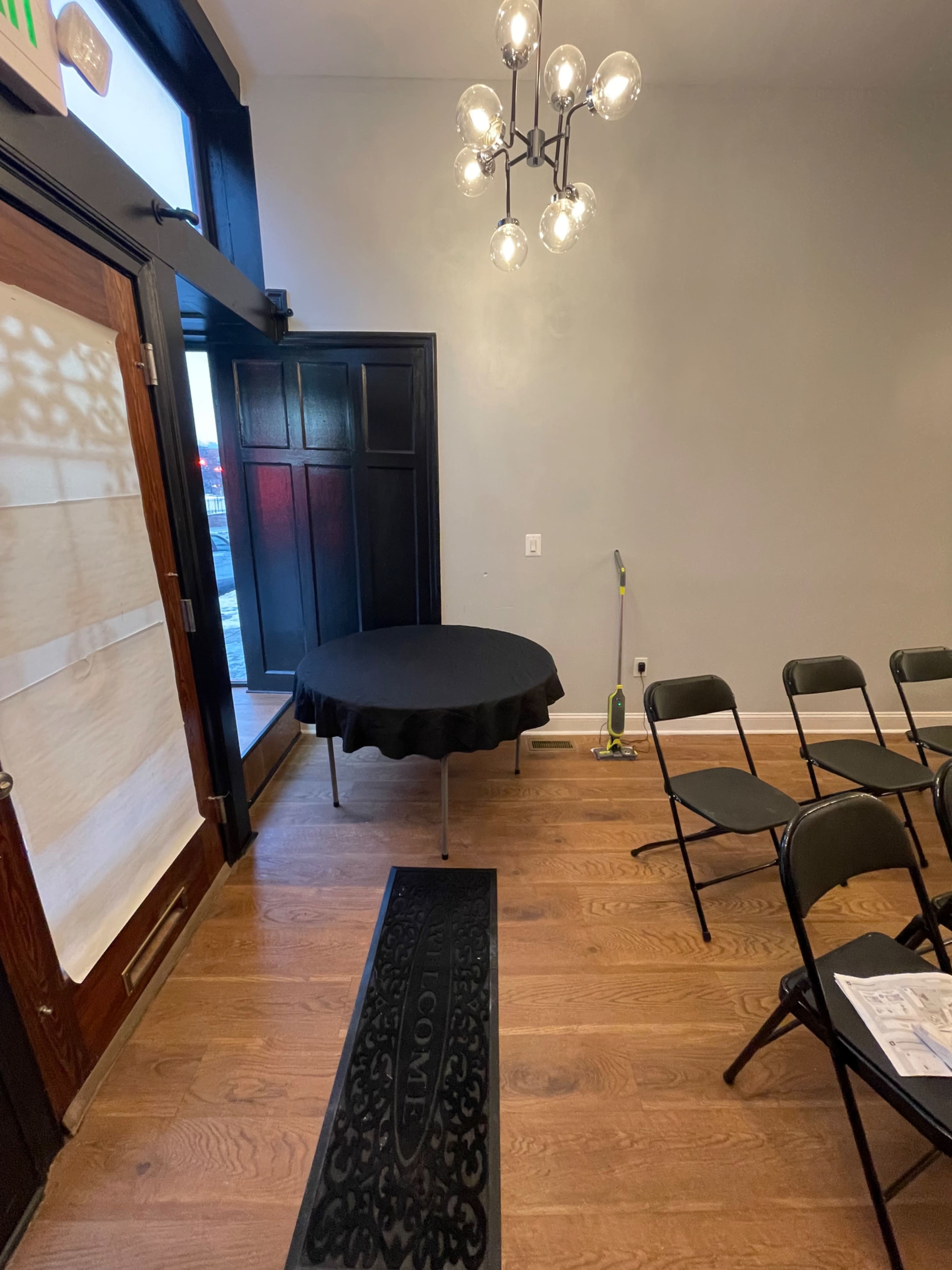 The image shows an indoor space featuring a round black table with a tablecloth, a wooden door, a chandelier, and folding chairs arranged in front of it.