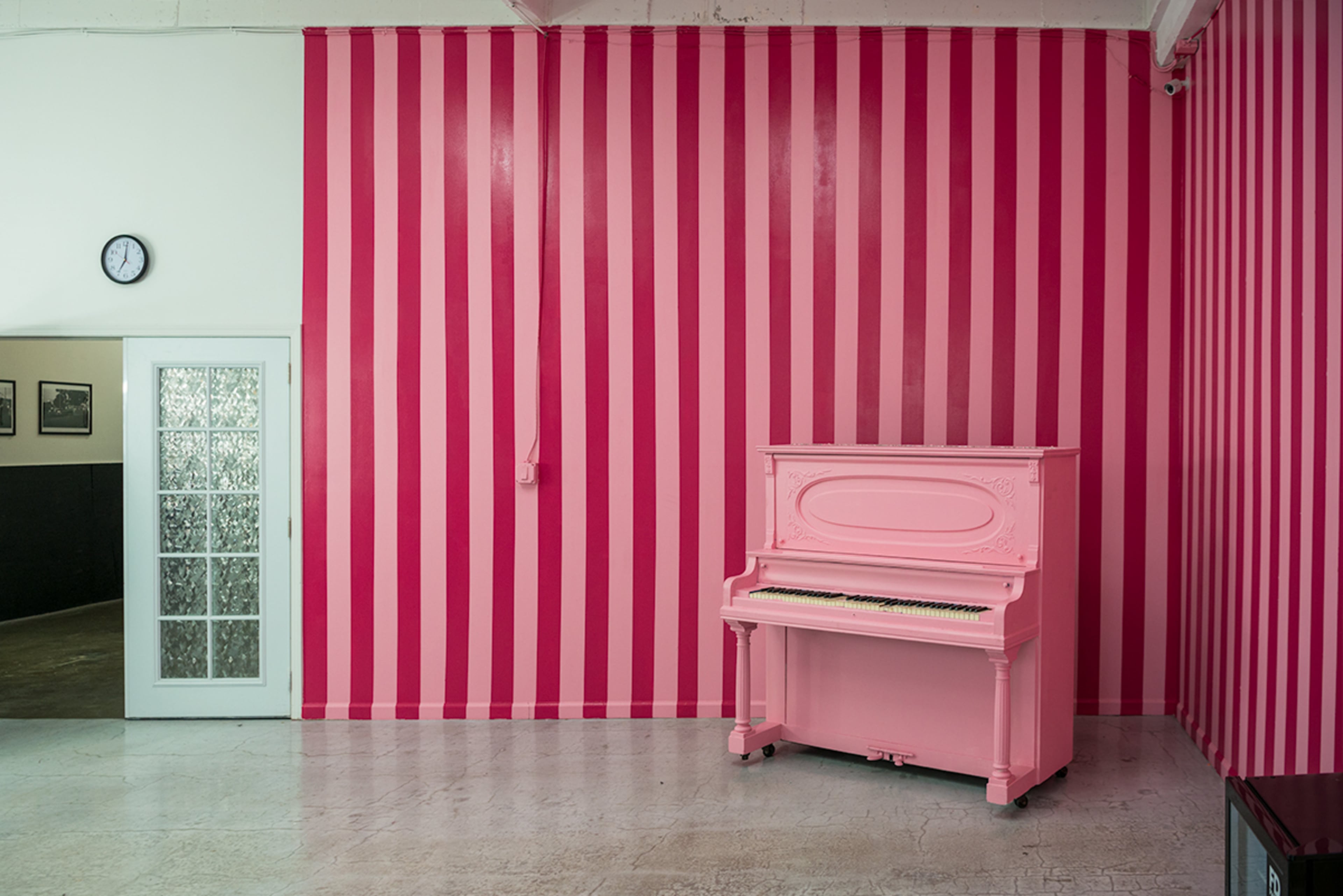 A pink piano stands against a wall decorated with vertical pink and white stripes in a room featuring a clock and a glass-paneled door.