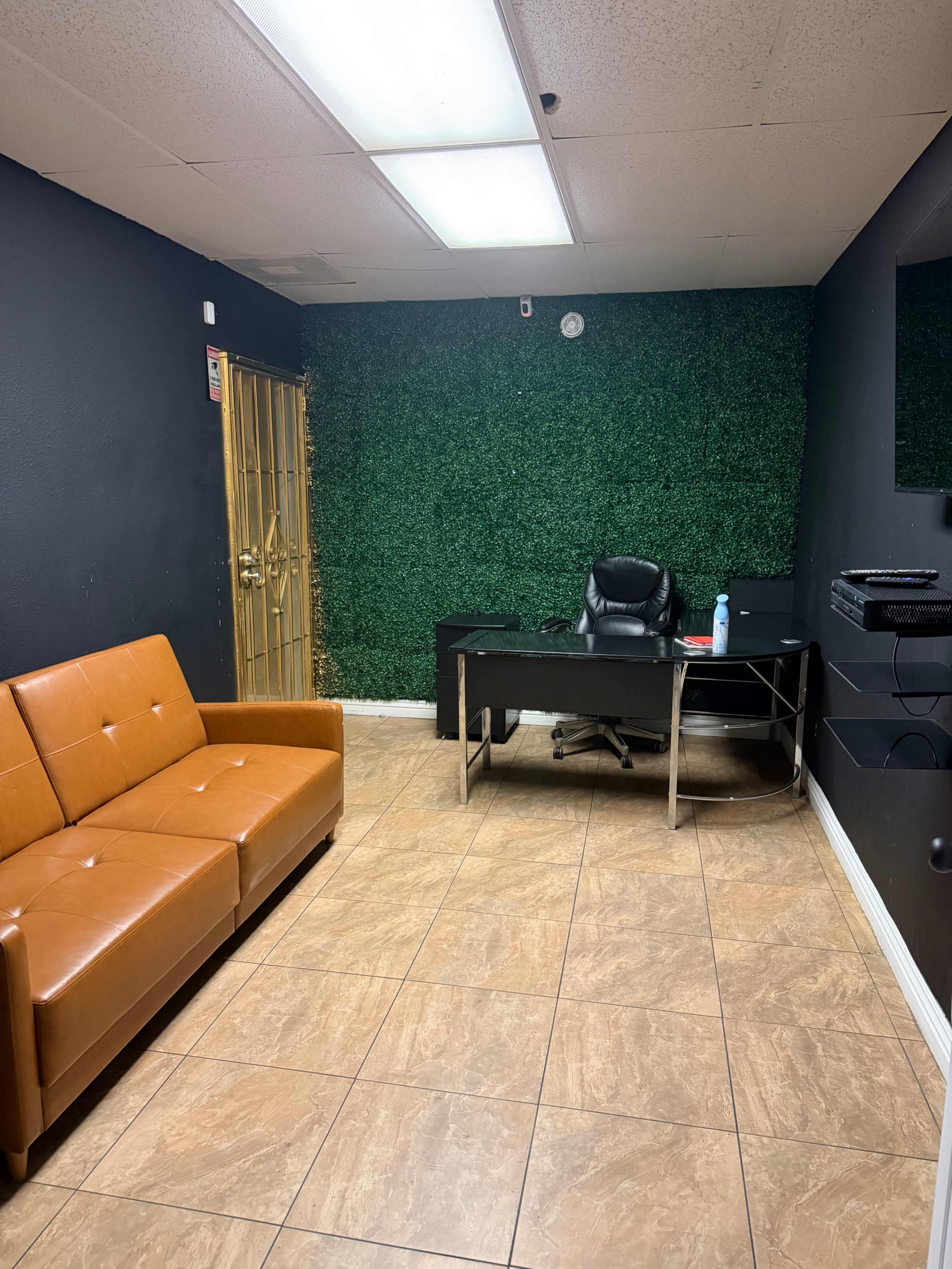 The image shows an office space with a brown sofa, a black desk, and a green hedge-like wall decoration.