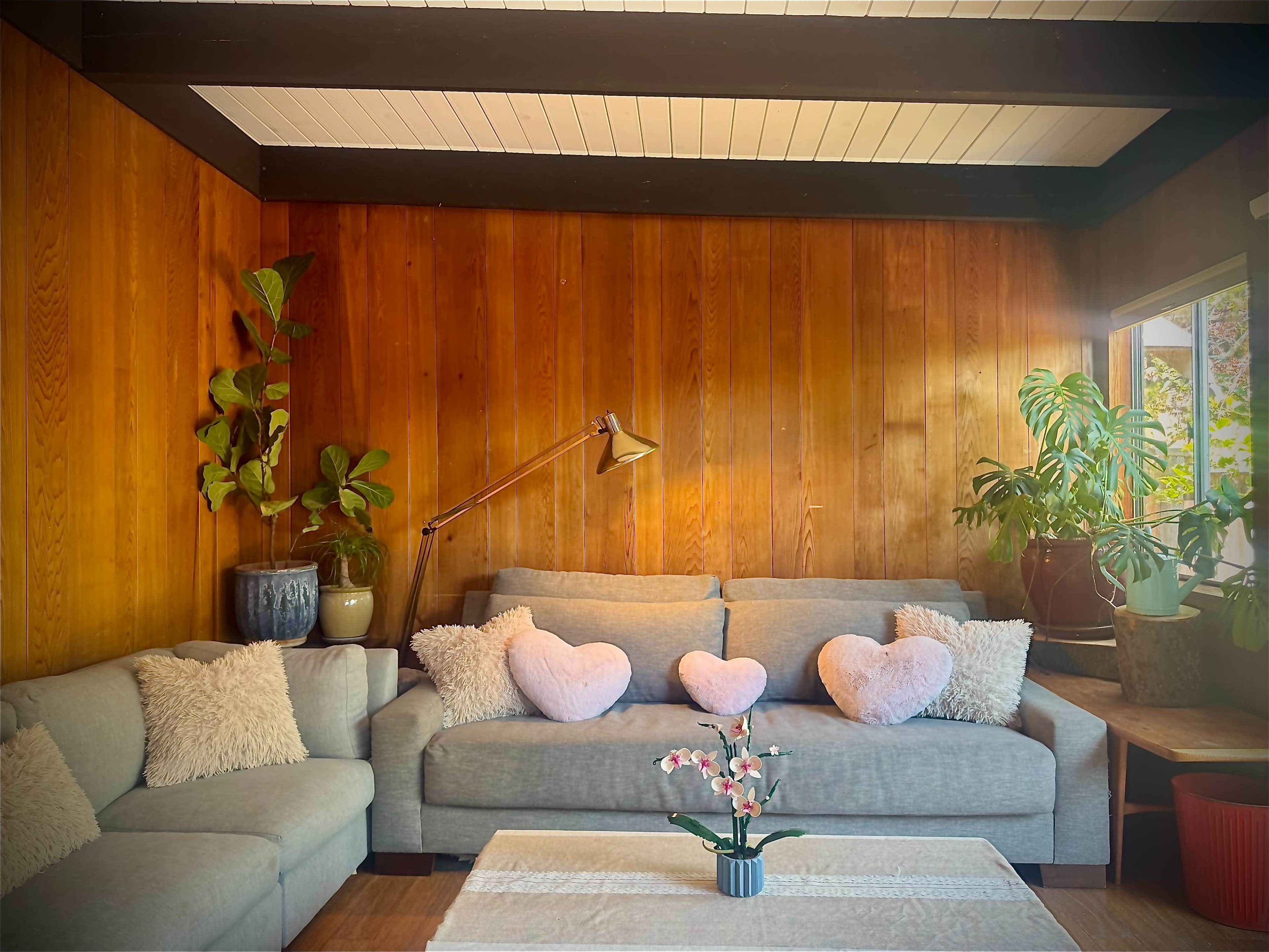 A modern living room features a light gray sofa with heart-shaped cushions, a coffee table, and potted plants against wooden-paneled walls.