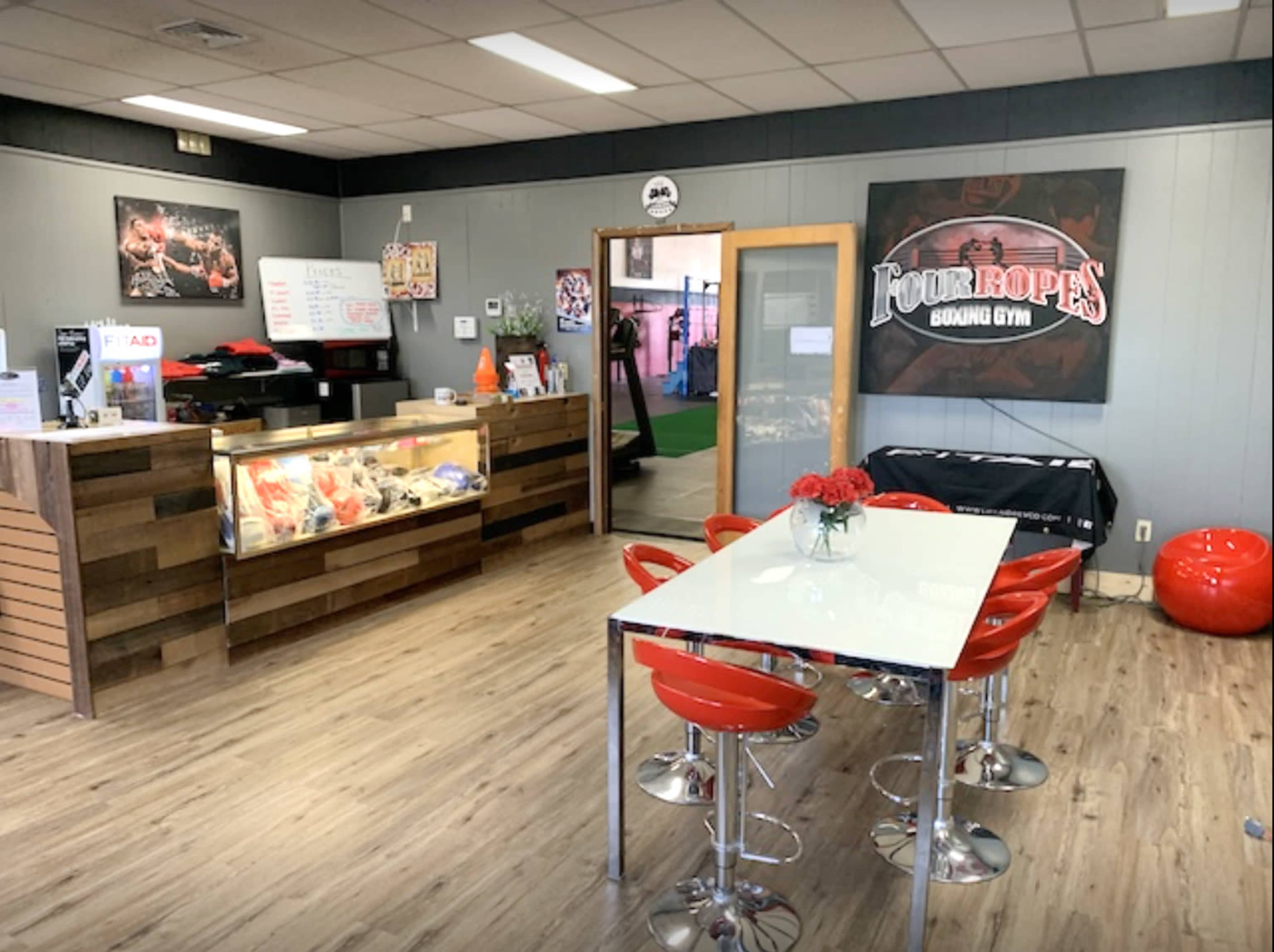 The image shows the interior of a boxing gym's reception area, featuring a front desk, a display case, a dining table with red chairs, and fitness-related decor on the walls.