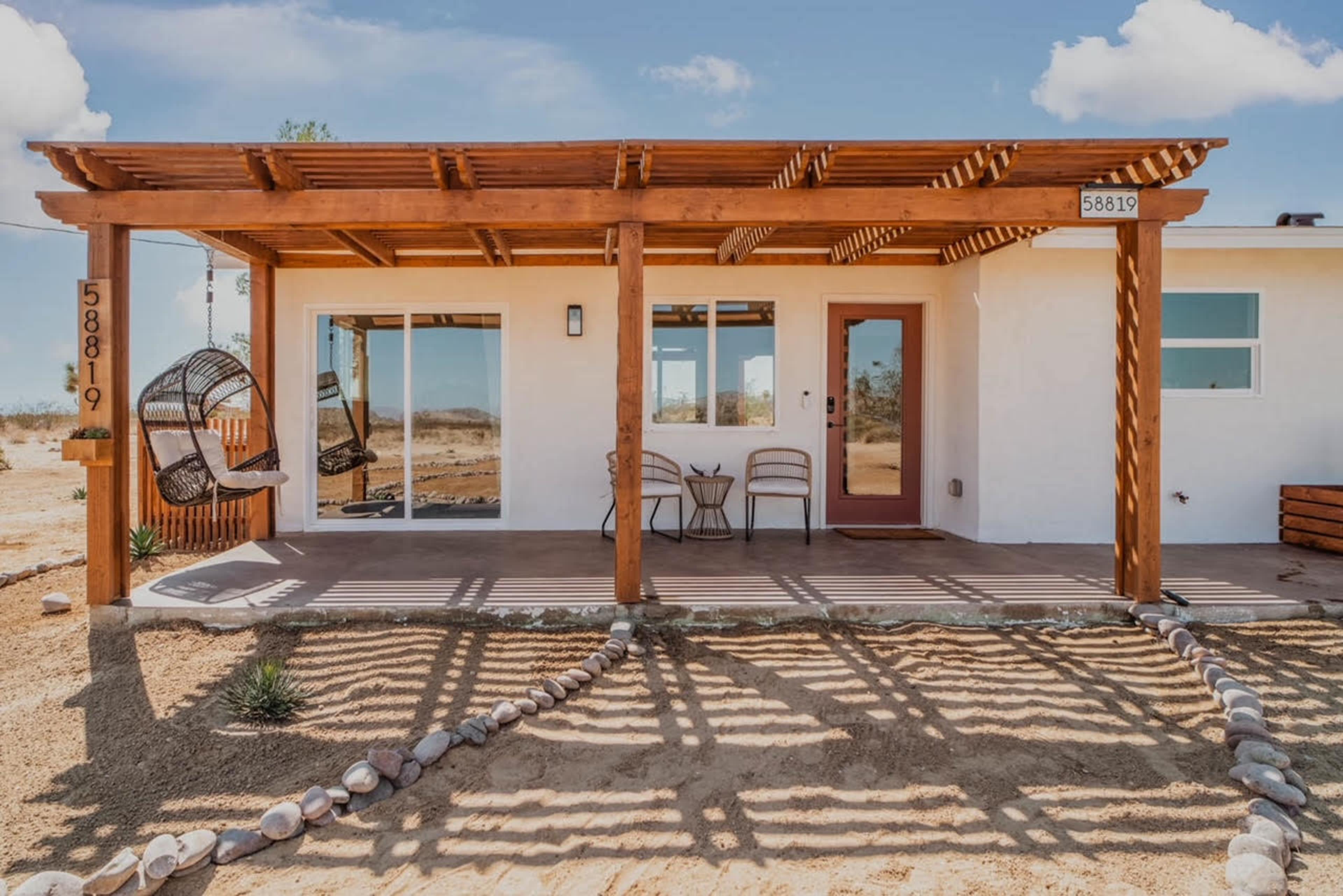 A modern one-story house with a wooden pergola, swinging chair, and two chairs on a sandy front porch.