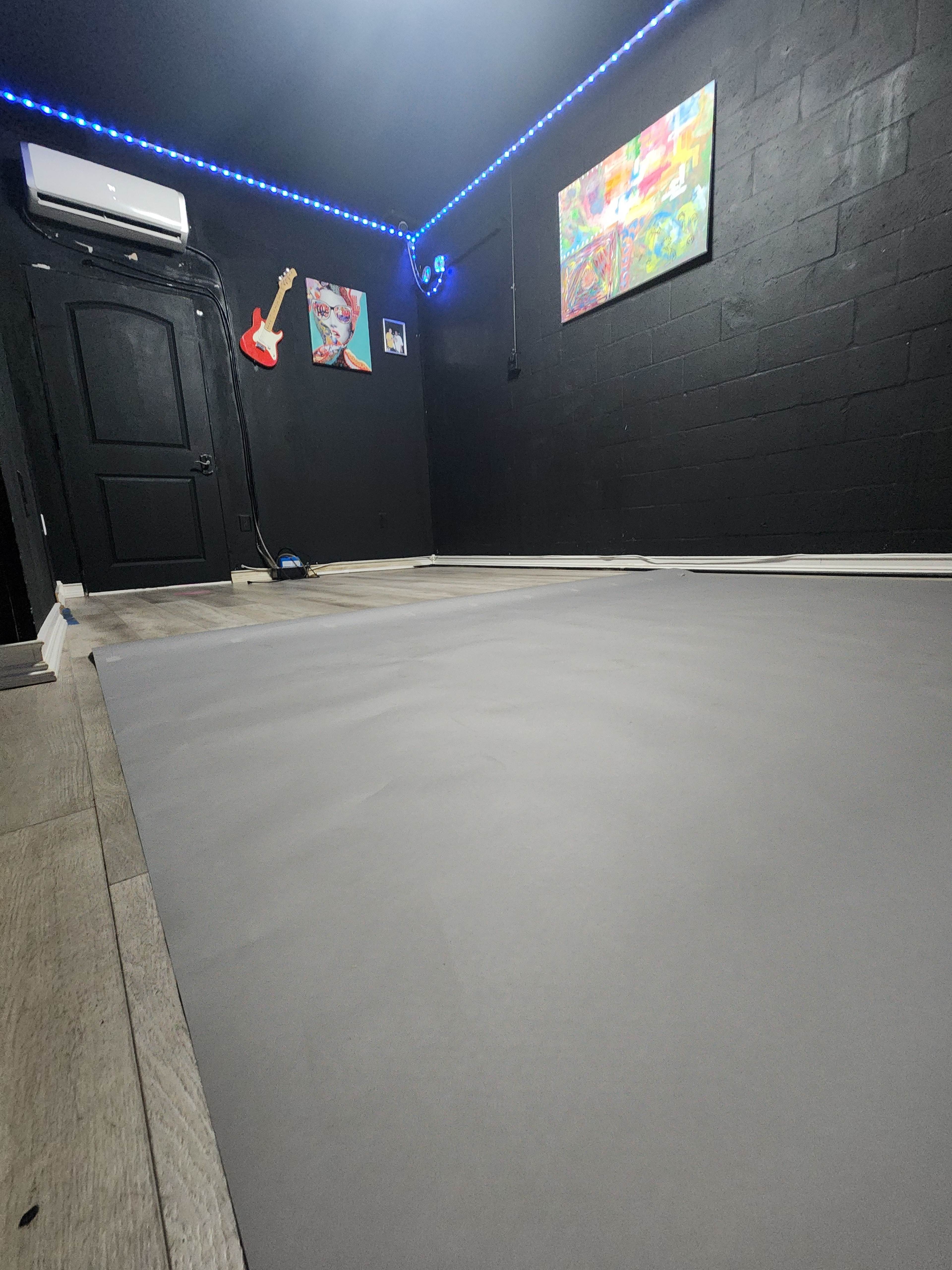 The image shows a spacious room with a dark black wall, a gray floor mat, colorful artwork hanging, and blue LED lights.