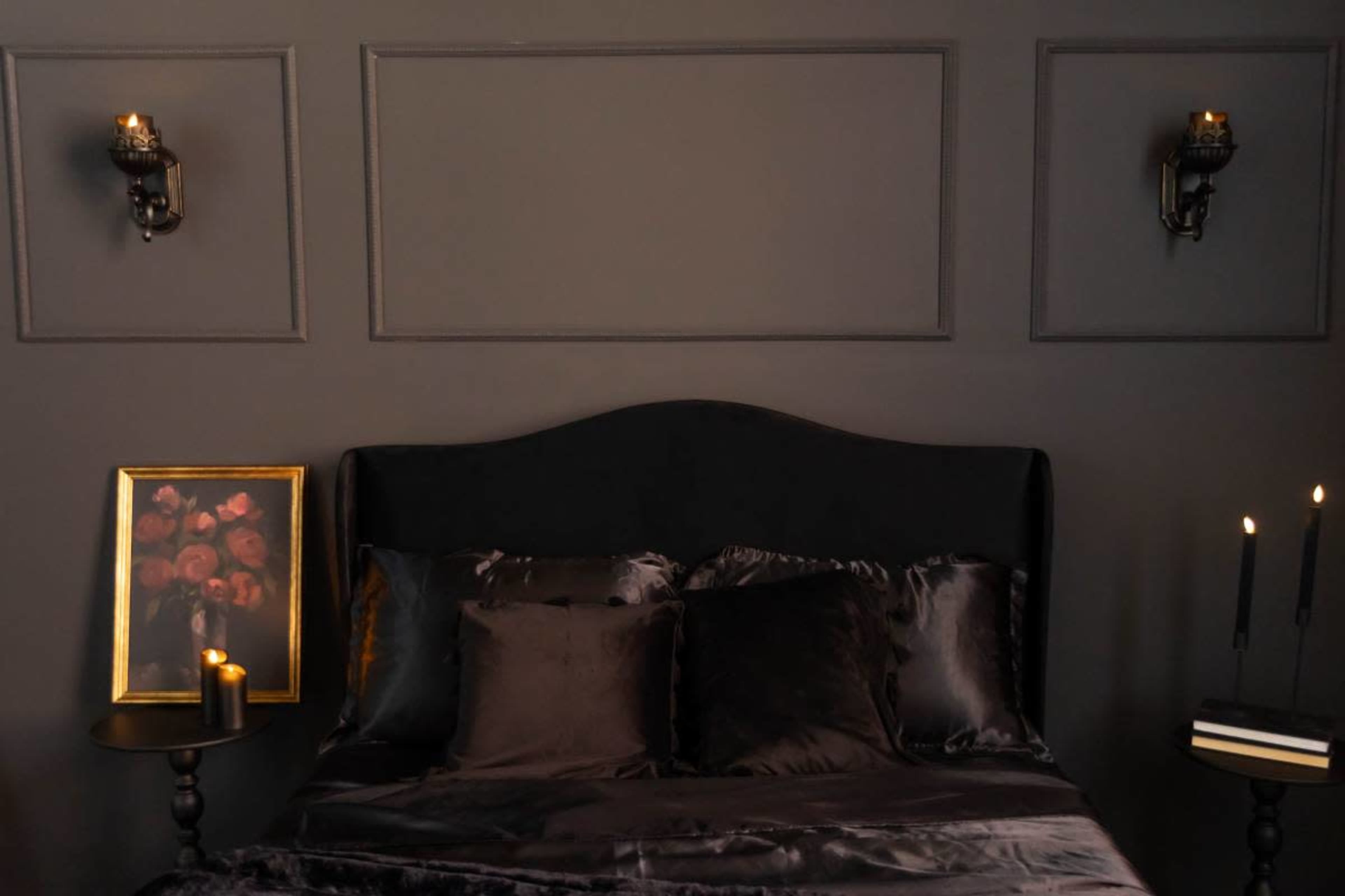The image shows a dark-colored bedroom with a black bed adorned with silky pillows, framed artwork of roses, and wall sconces casting a soft glow.