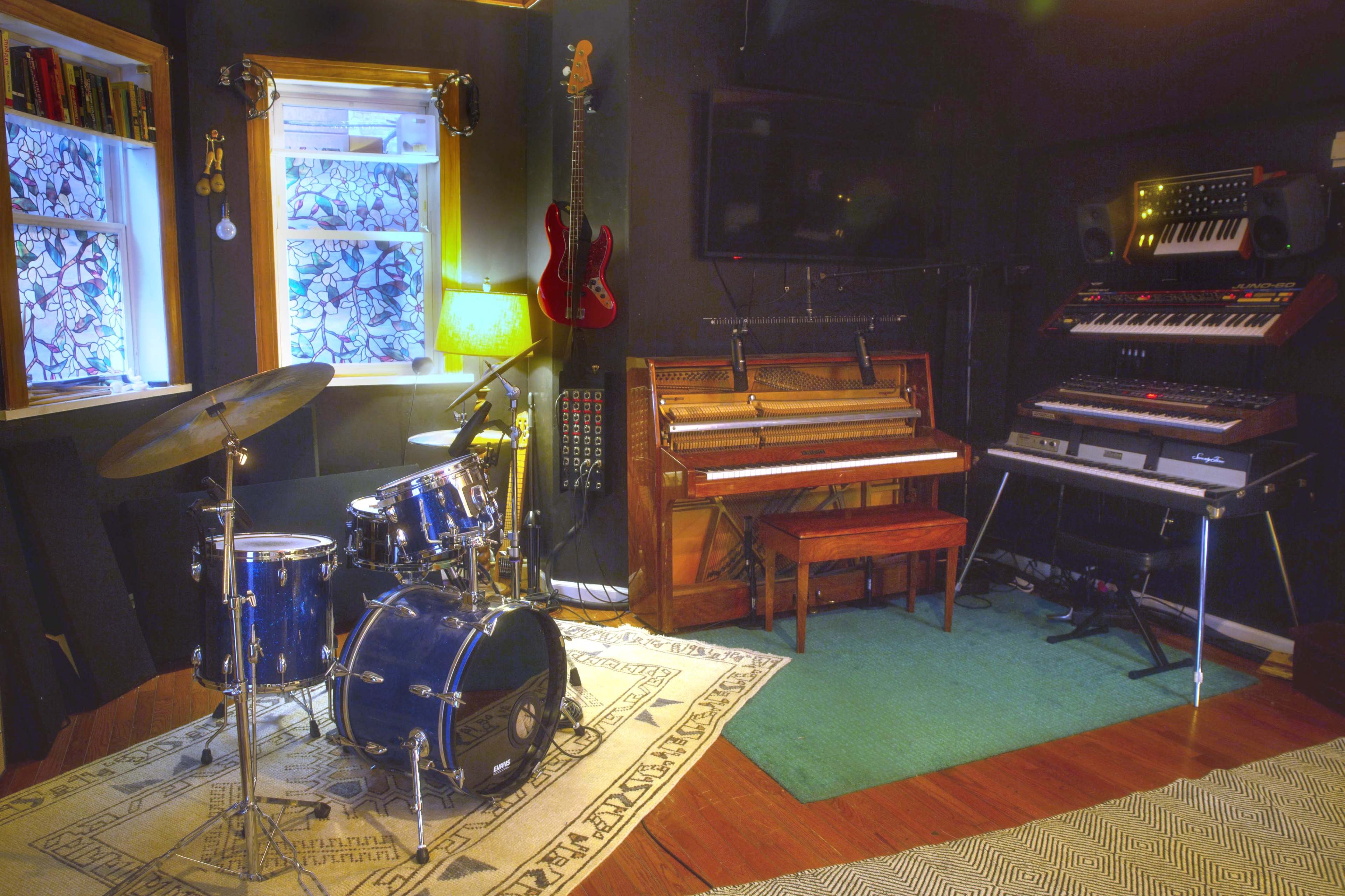 A music studio featuring a drum set, an acoustic piano, and electronic keyboards arranged against a dark wall, with windows providing natural light.