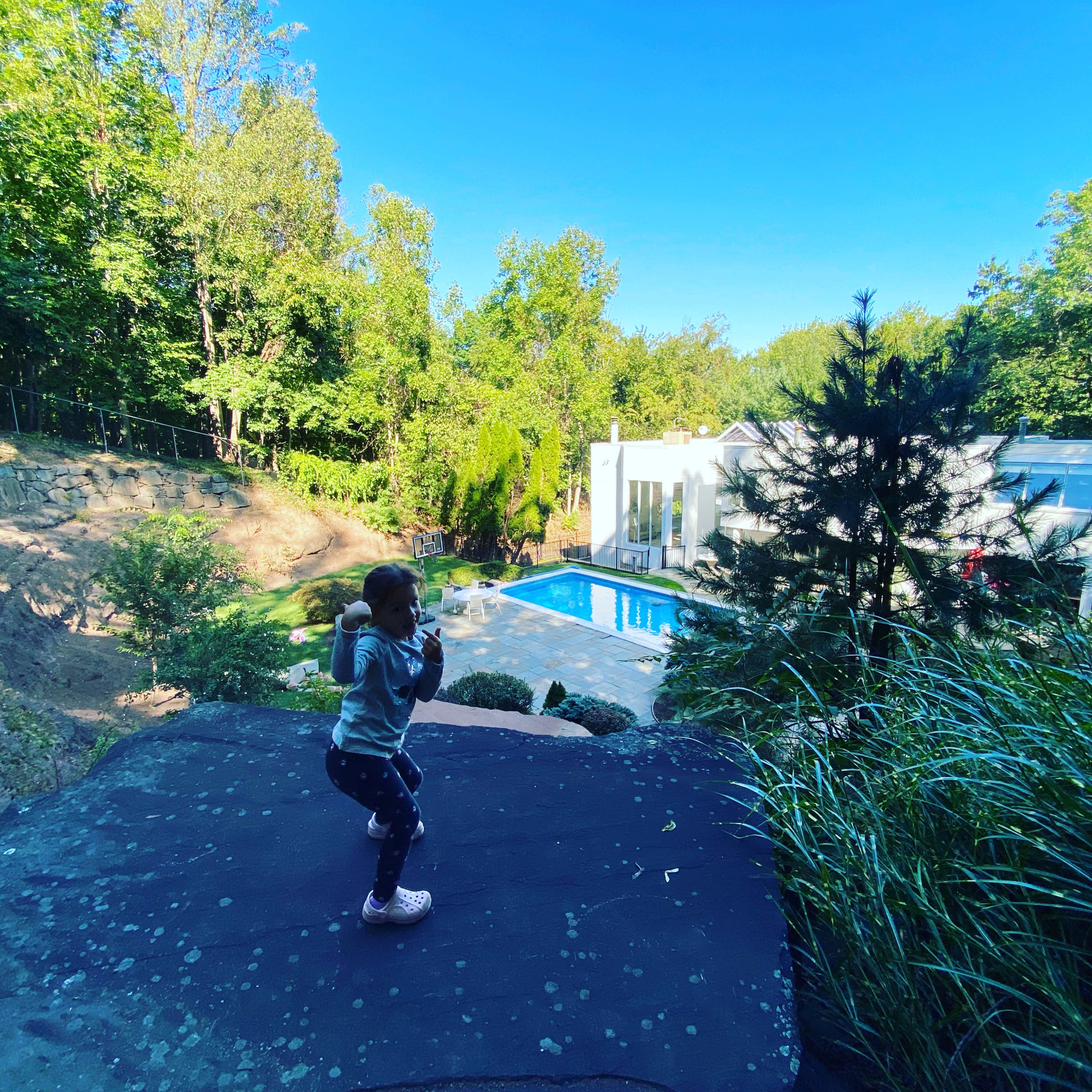 A child stands on a rock outcrop overlooking a modern house with a pool surrounded by greenery.