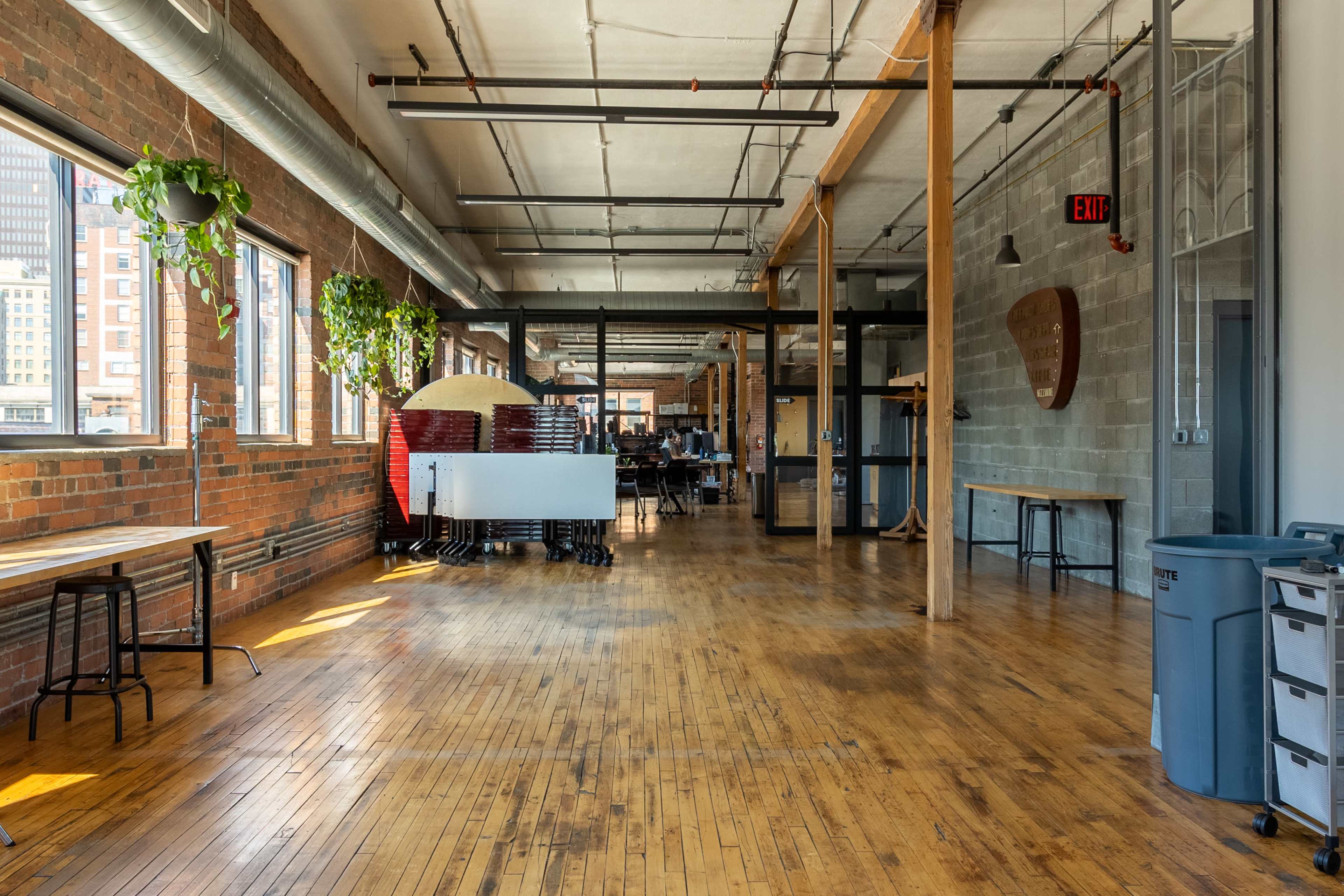 A spacious, industrial-style room with wooden floors, large windows, and exposed brick walls, featuring furniture and plants along the walls.