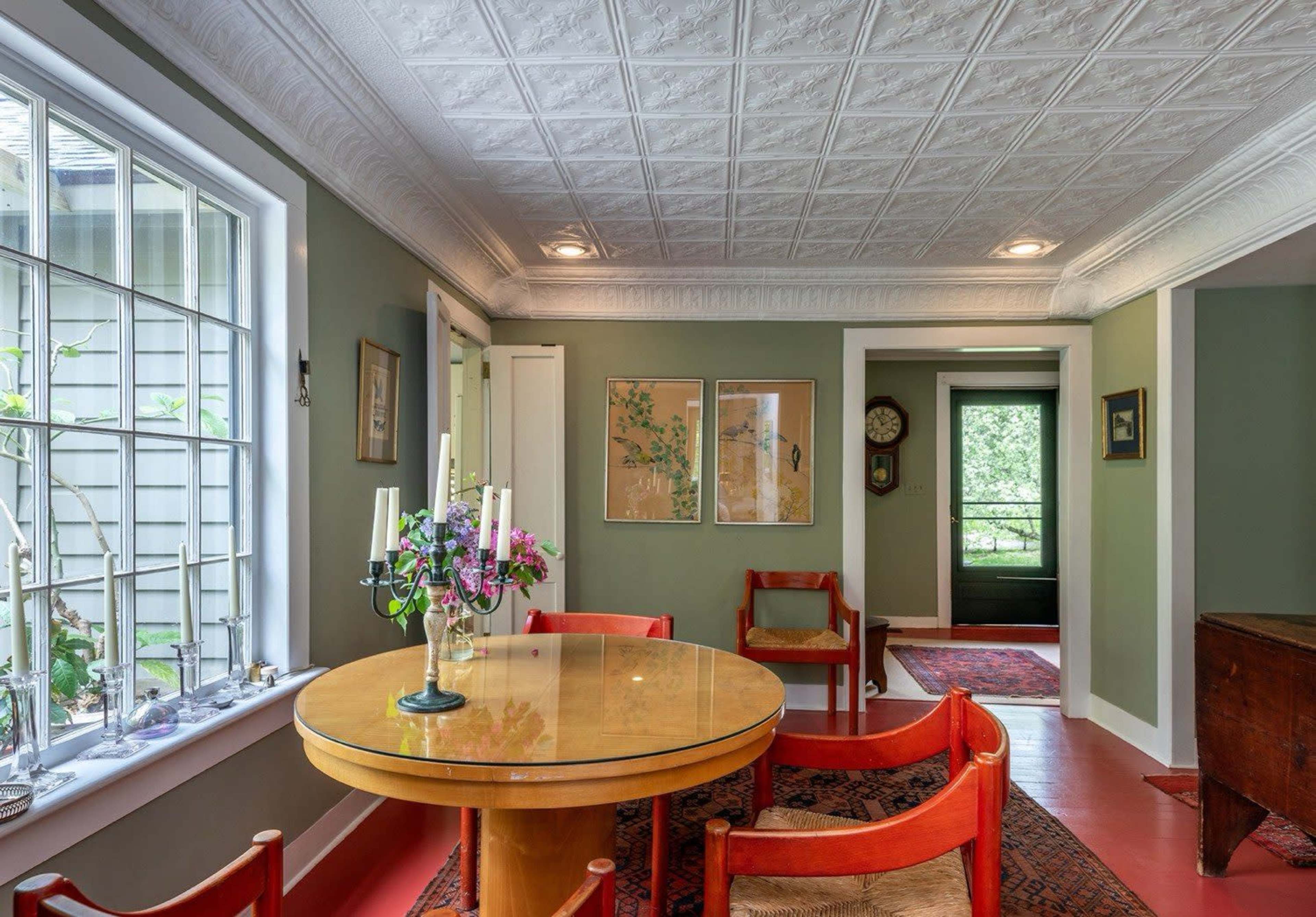 The image shows a dining area with a round table surrounded by red chairs, floral decorations, and a view of a hallway leading to another room.