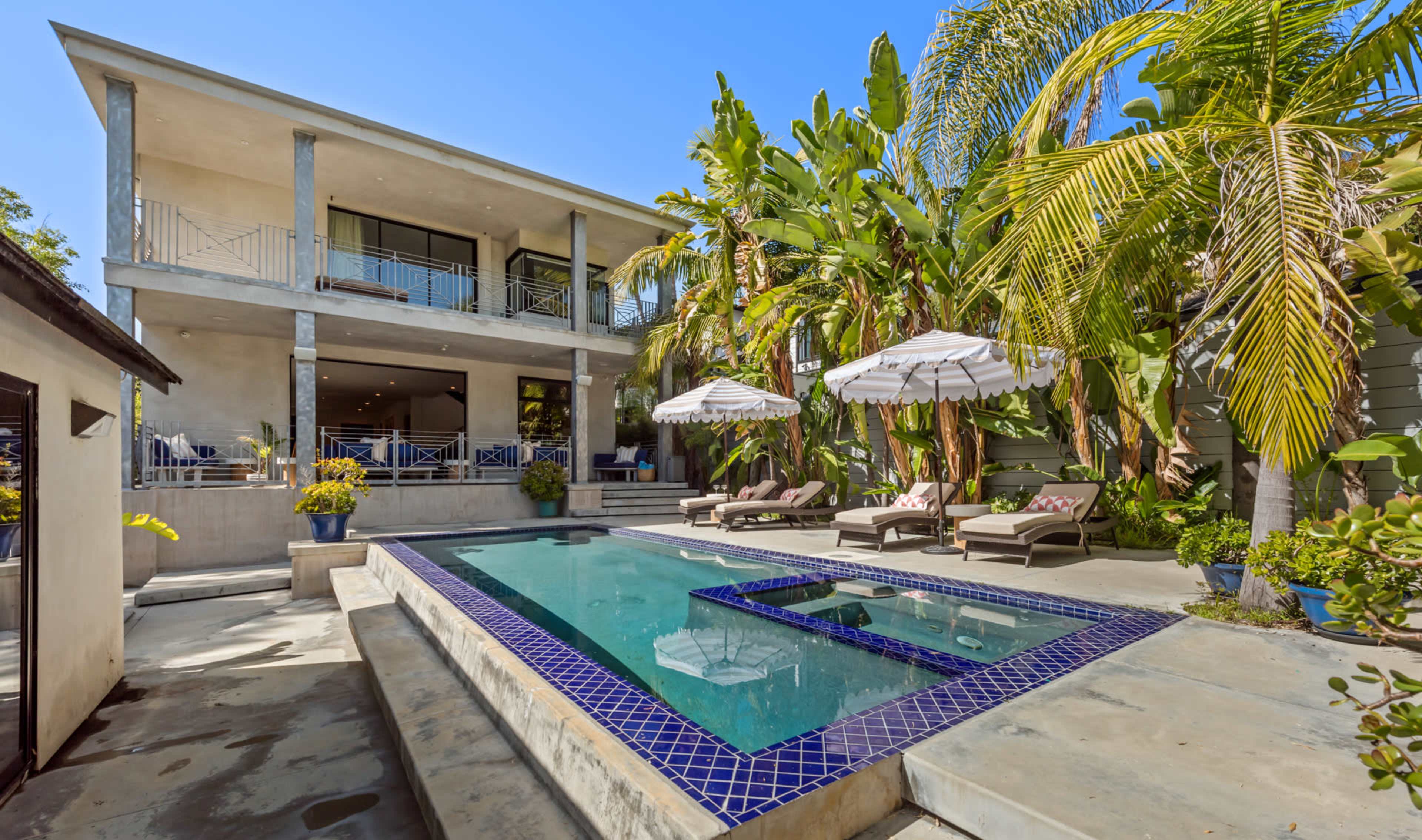 The image shows a modern two-story house with a swimming pool surrounded by lounge chairs and palm trees.