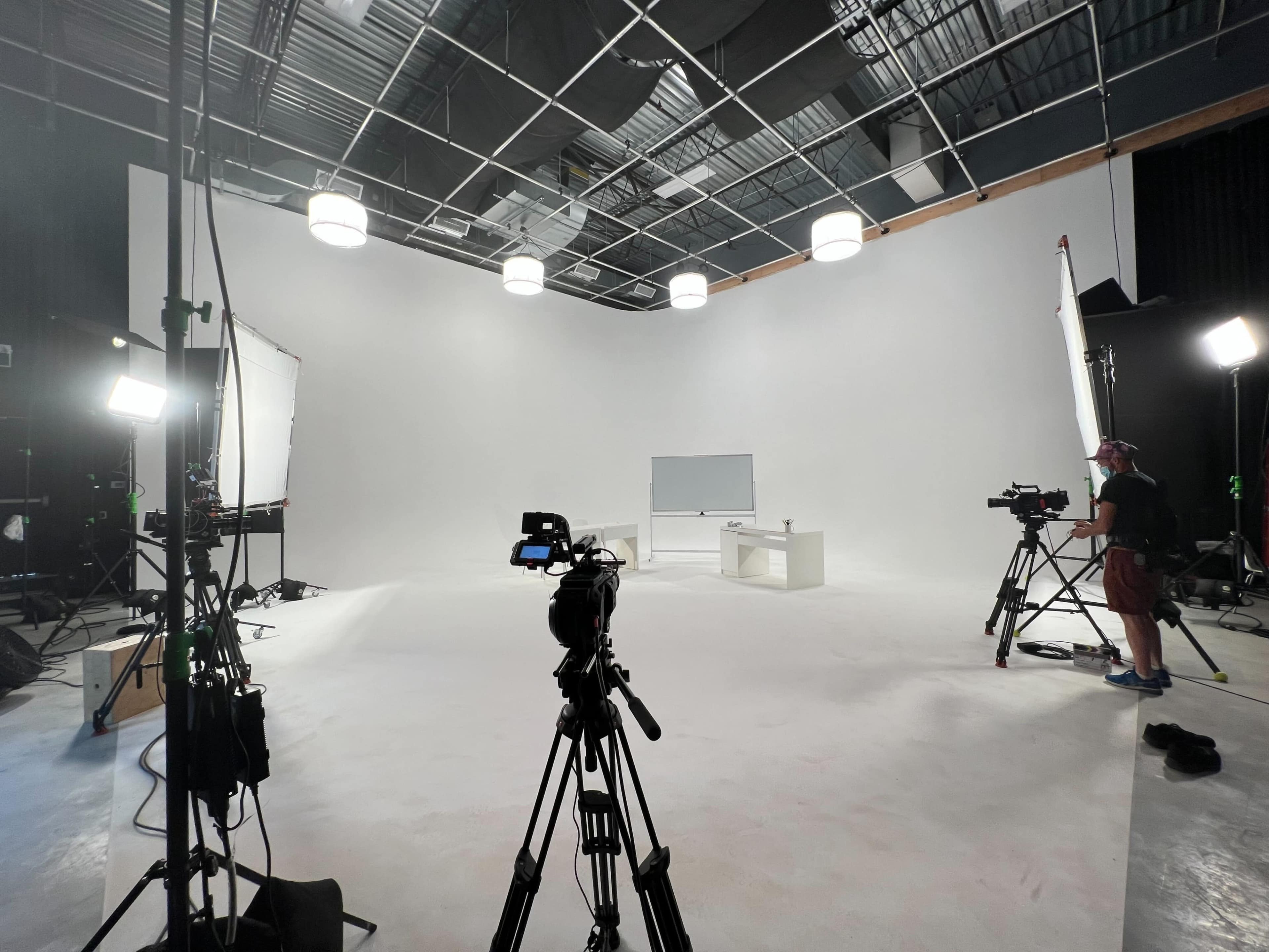 A film studio setup features a white backdrop, two cameras on tripods, a whiteboard, and several lighting fixtures.