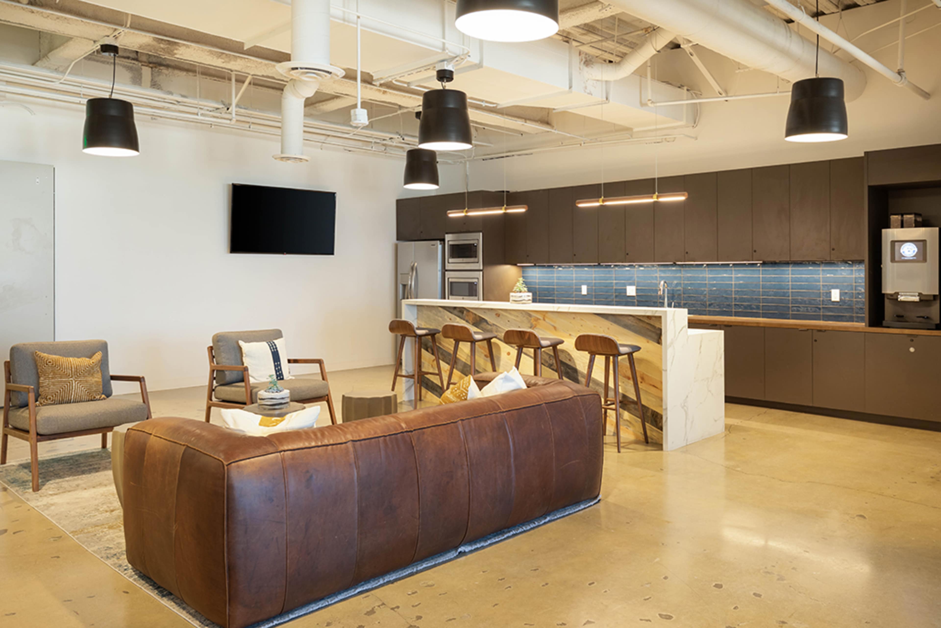A modern office break room featuring a leather sofa, two armchairs, a kitchen bar with stools, and contemporary lighting fixtures.