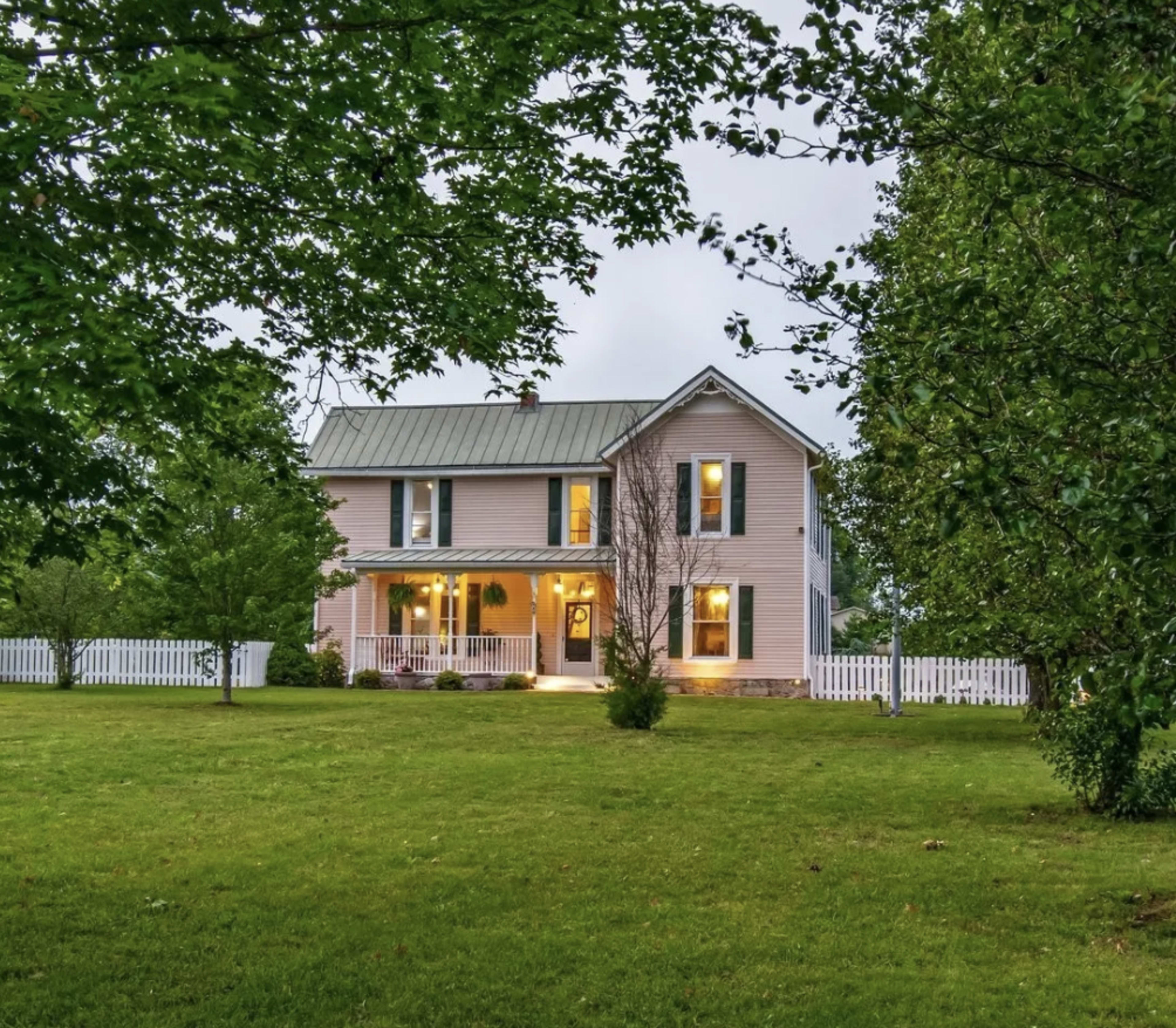 A two-story pink house with a metal roof is surrounded by a green lawn and trees, featuring a white picket fence in the front.