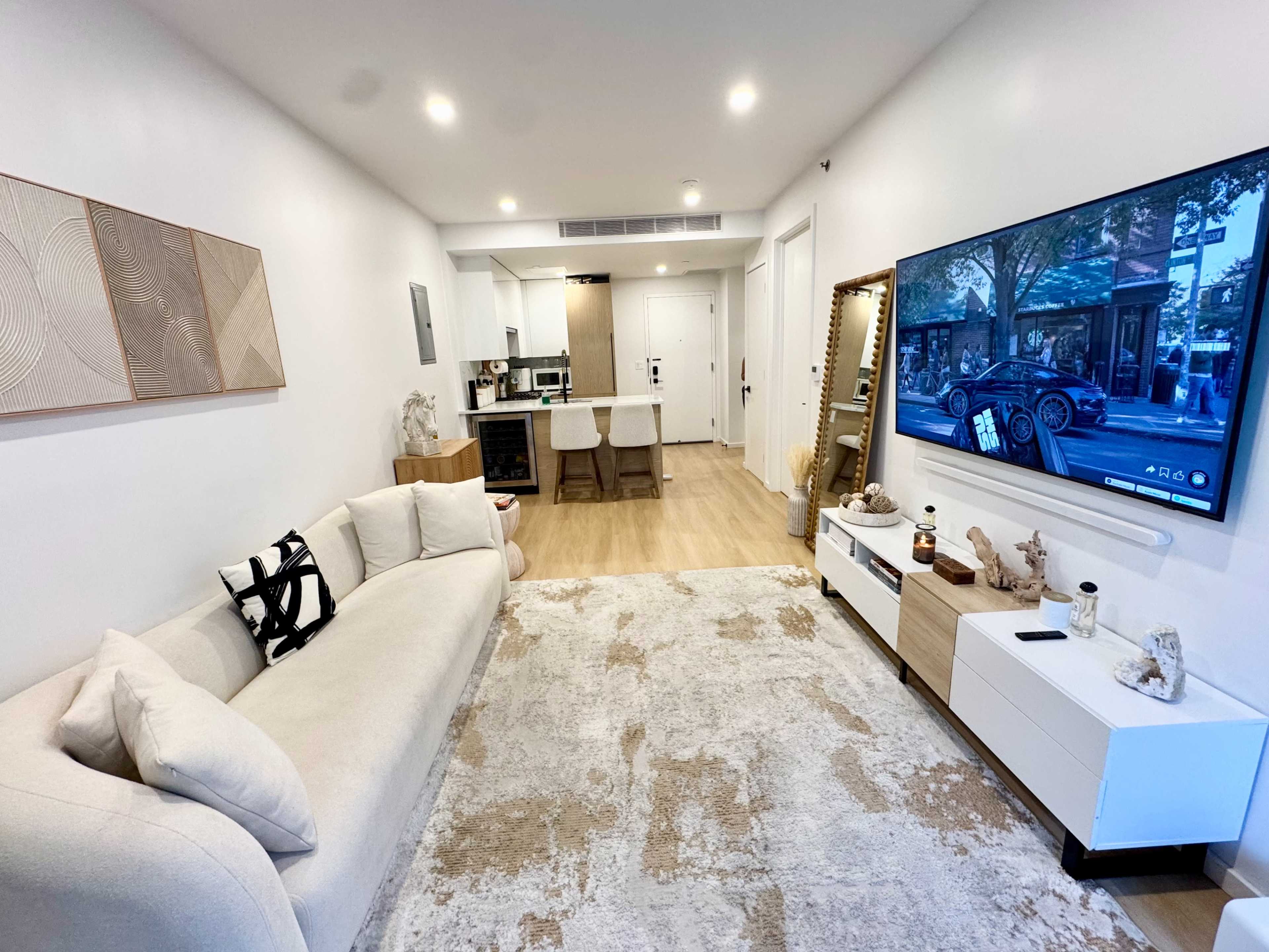 The image shows a modern living room featuring a light-colored sofa, a large television mounted on the wall, and a dining area visible in the background.
