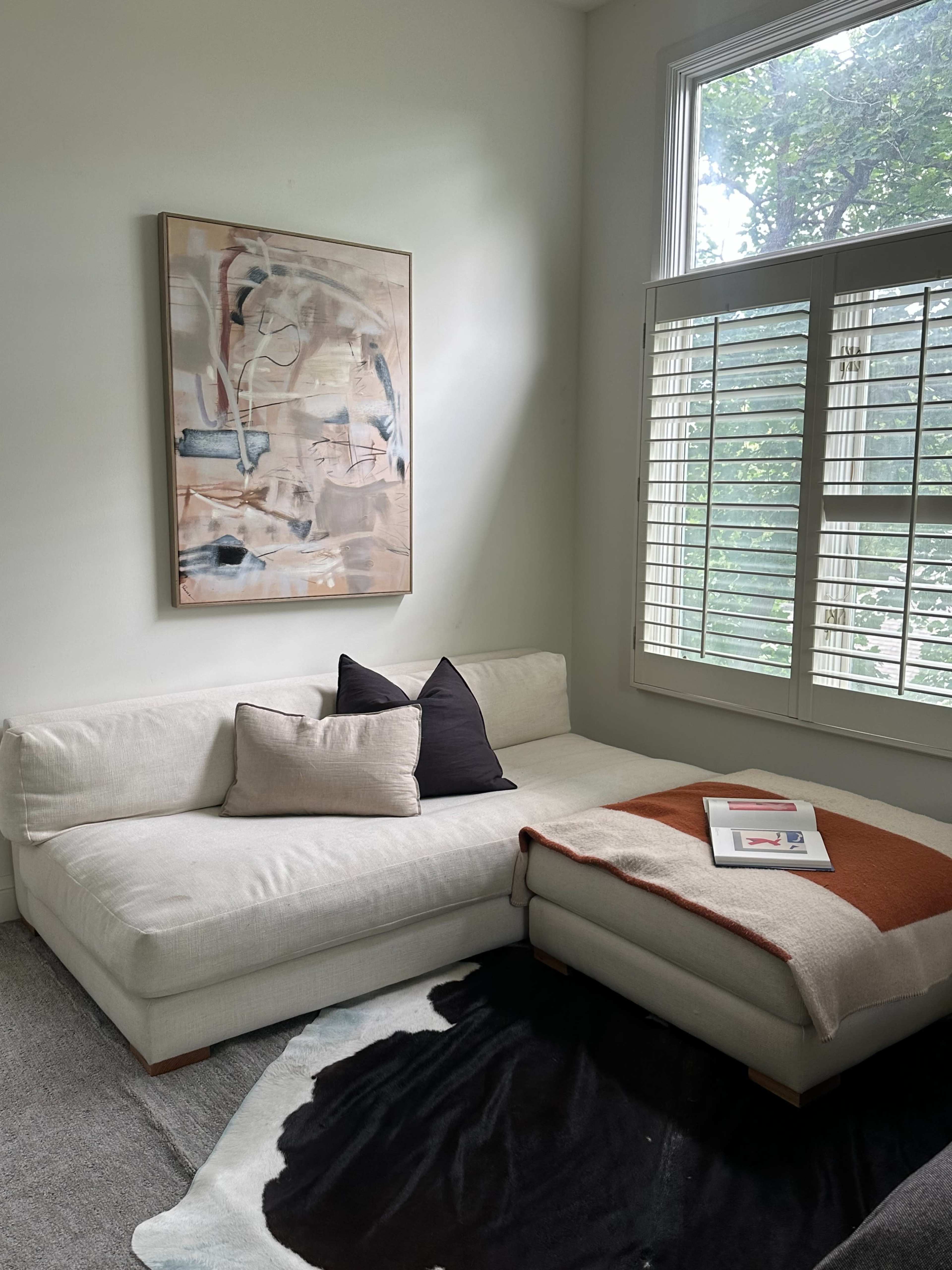 A corner of a living room features a light-colored sectional sofa with a black and white throw blanket, a small stack of magazines on the ottoman, and a large abstract painting above the sofa.