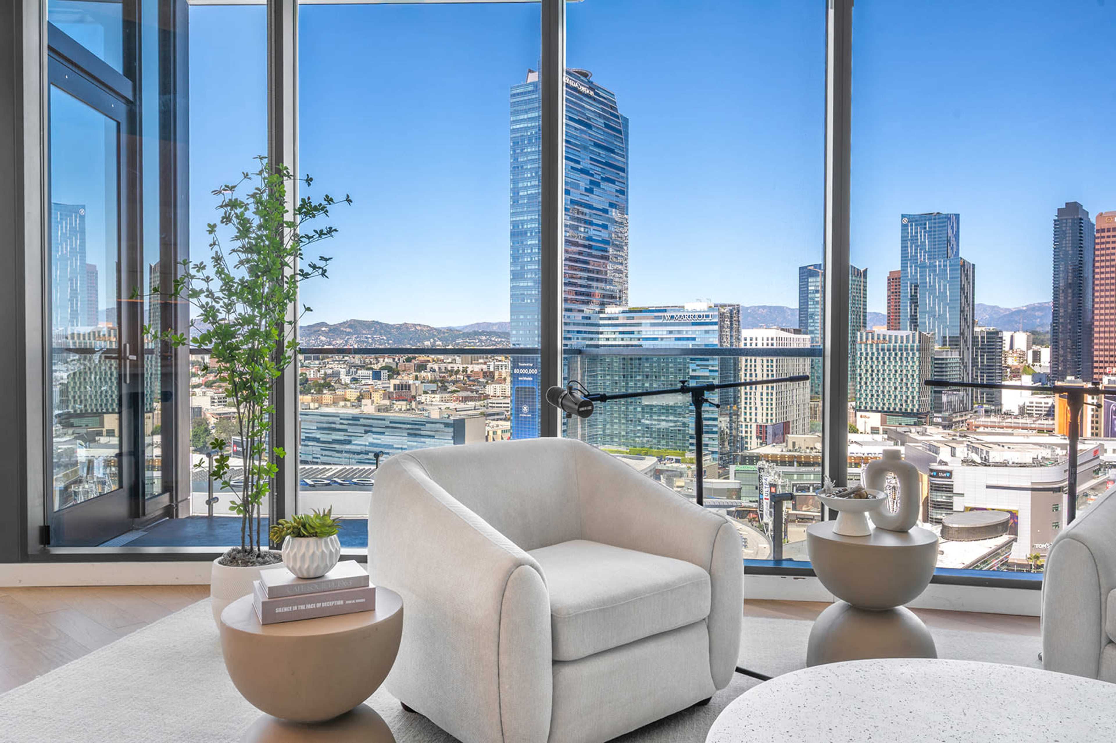 A modern living room features light-colored furniture and large windows showcasing a view of a city skyline with tall buildings and mountains in the background.