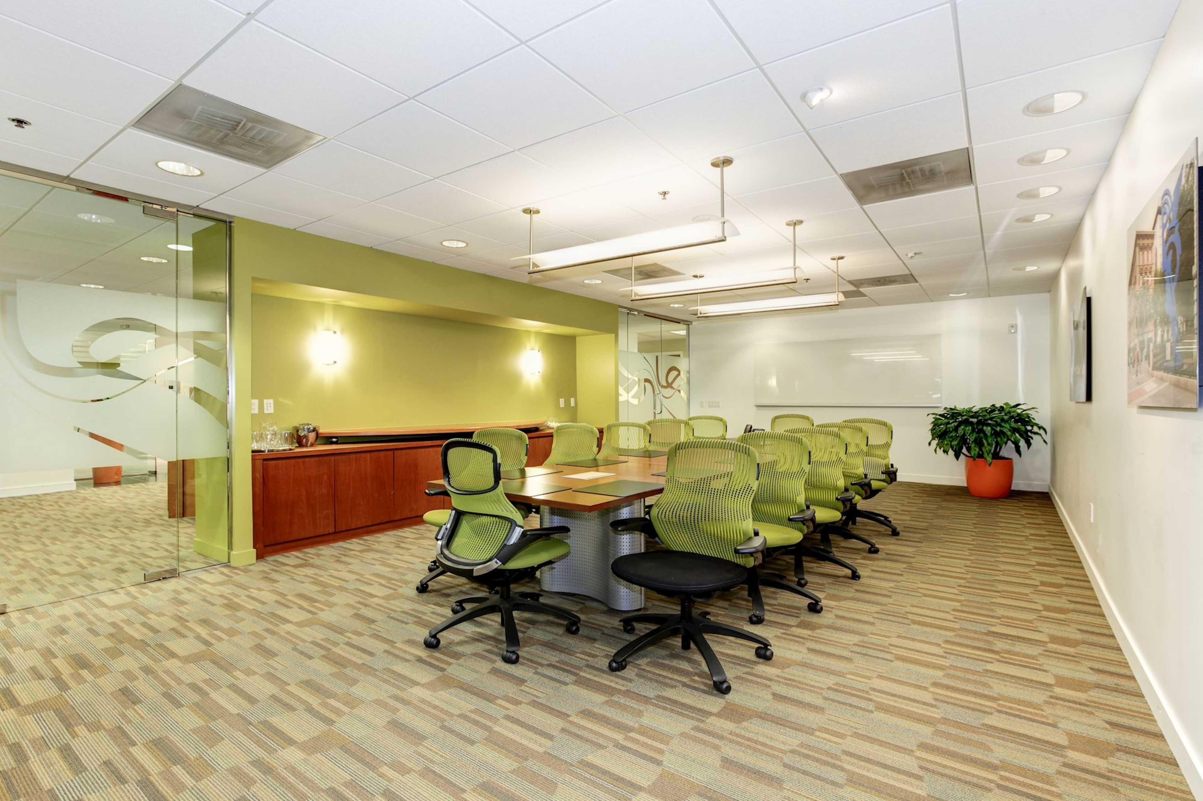 A modern conference room features a long table surrounded by ergonomic green chairs, with glass walls and a decorative plant in the corner.