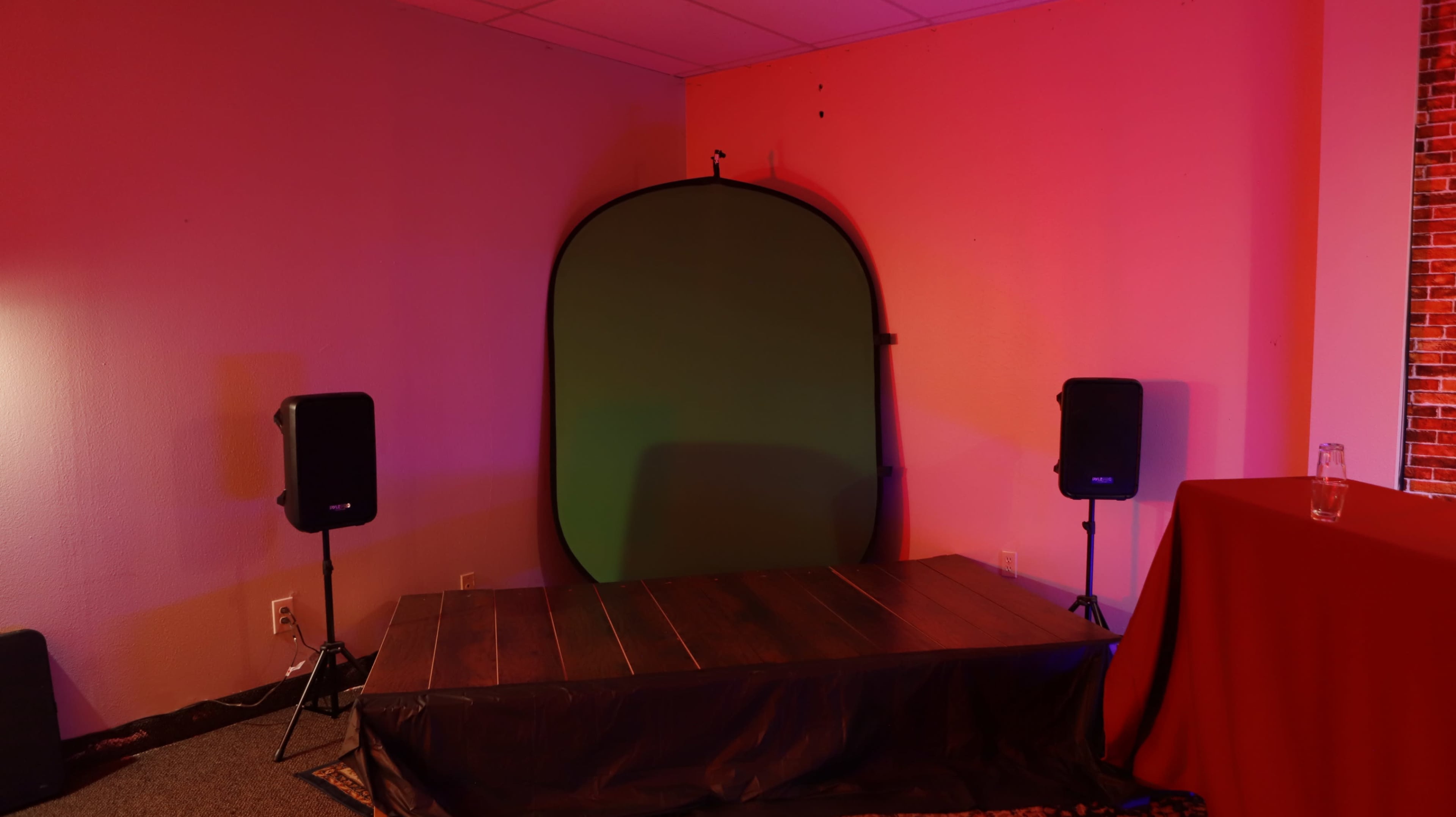 The image shows a small stage area with a green backdrop, two speakers on either side, and a table in the corner, all illuminated by colorful lighting.