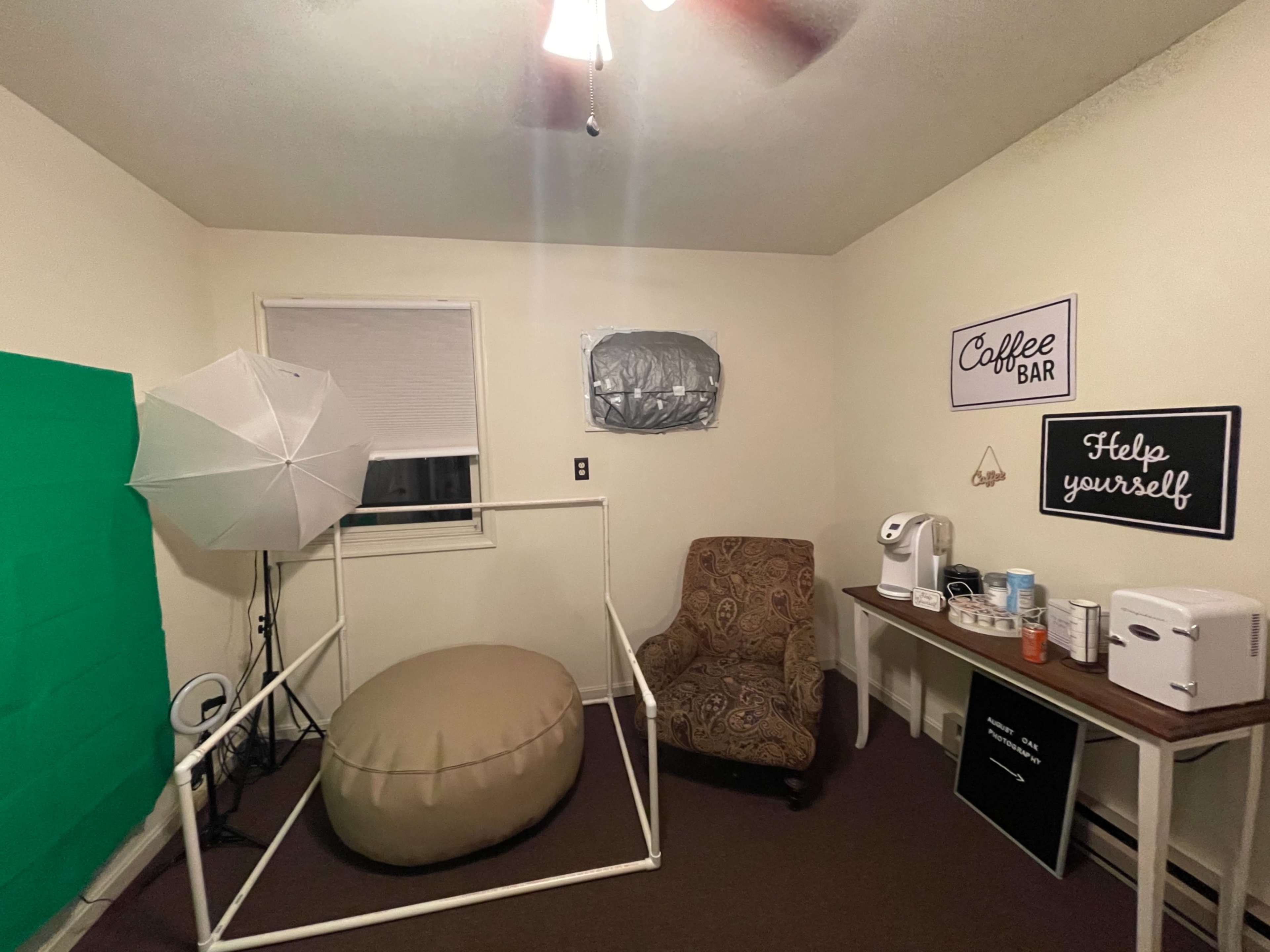 A small room with a light green backdrop, a large bean bag, a brown chair, a table with a coffee station, and softbox lighting equipment.