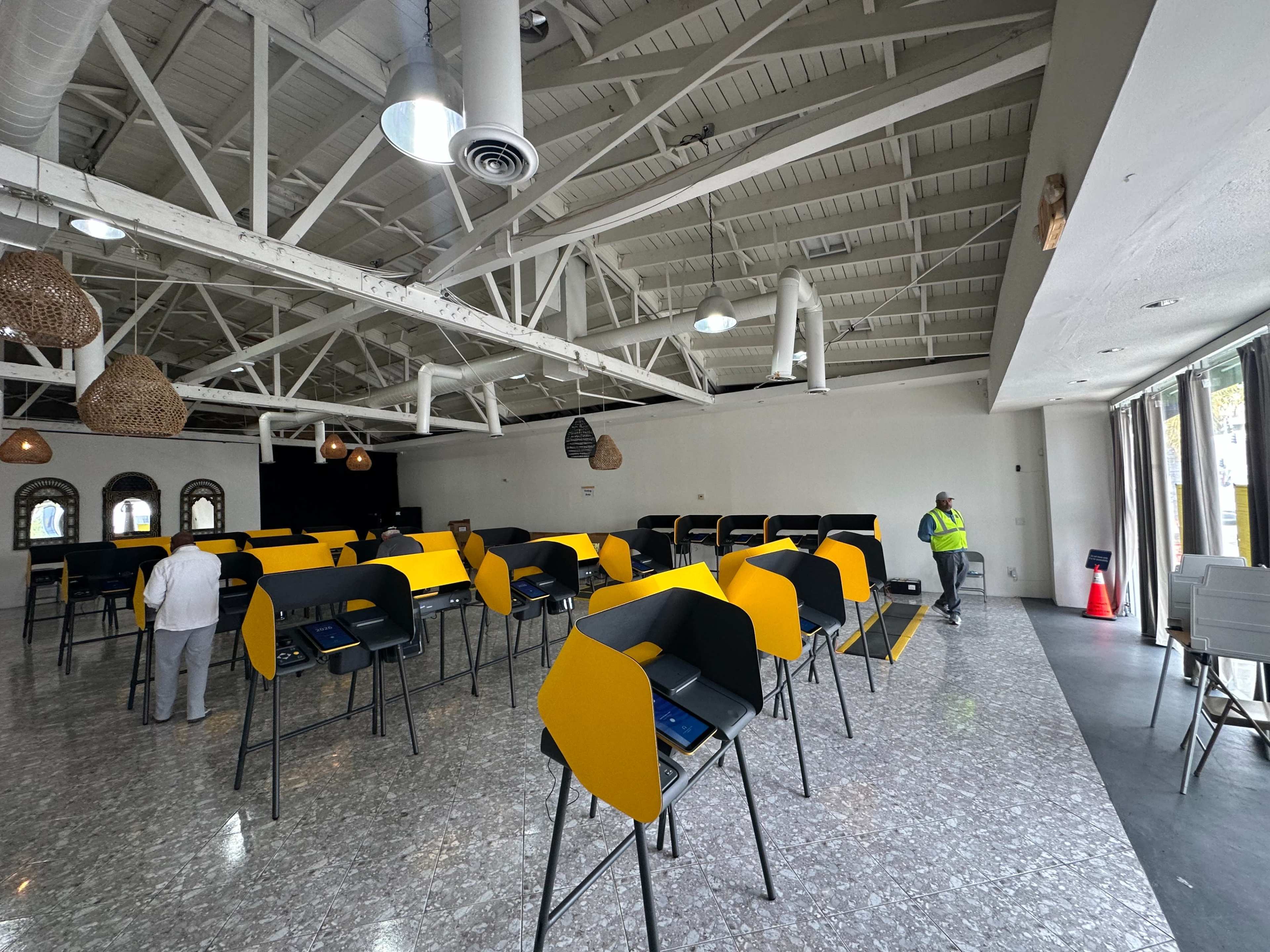 The image shows an empty polling station with multiple voting booths arranged in a spacious room featuring a high ceiling.