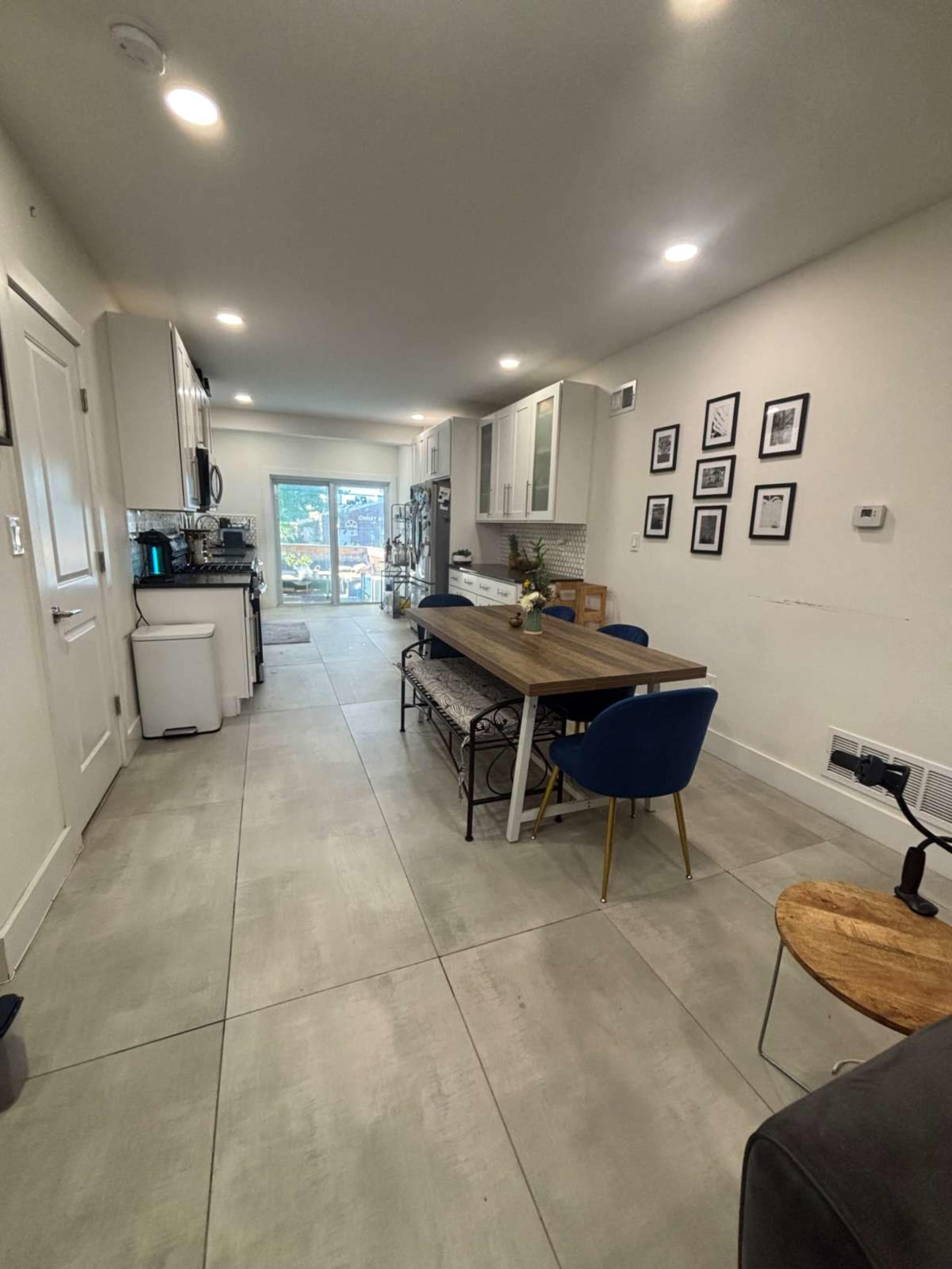 The image shows a modern kitchen and dining area with a long table and blue chairs, leading to a patio door that opens to an outdoor space.