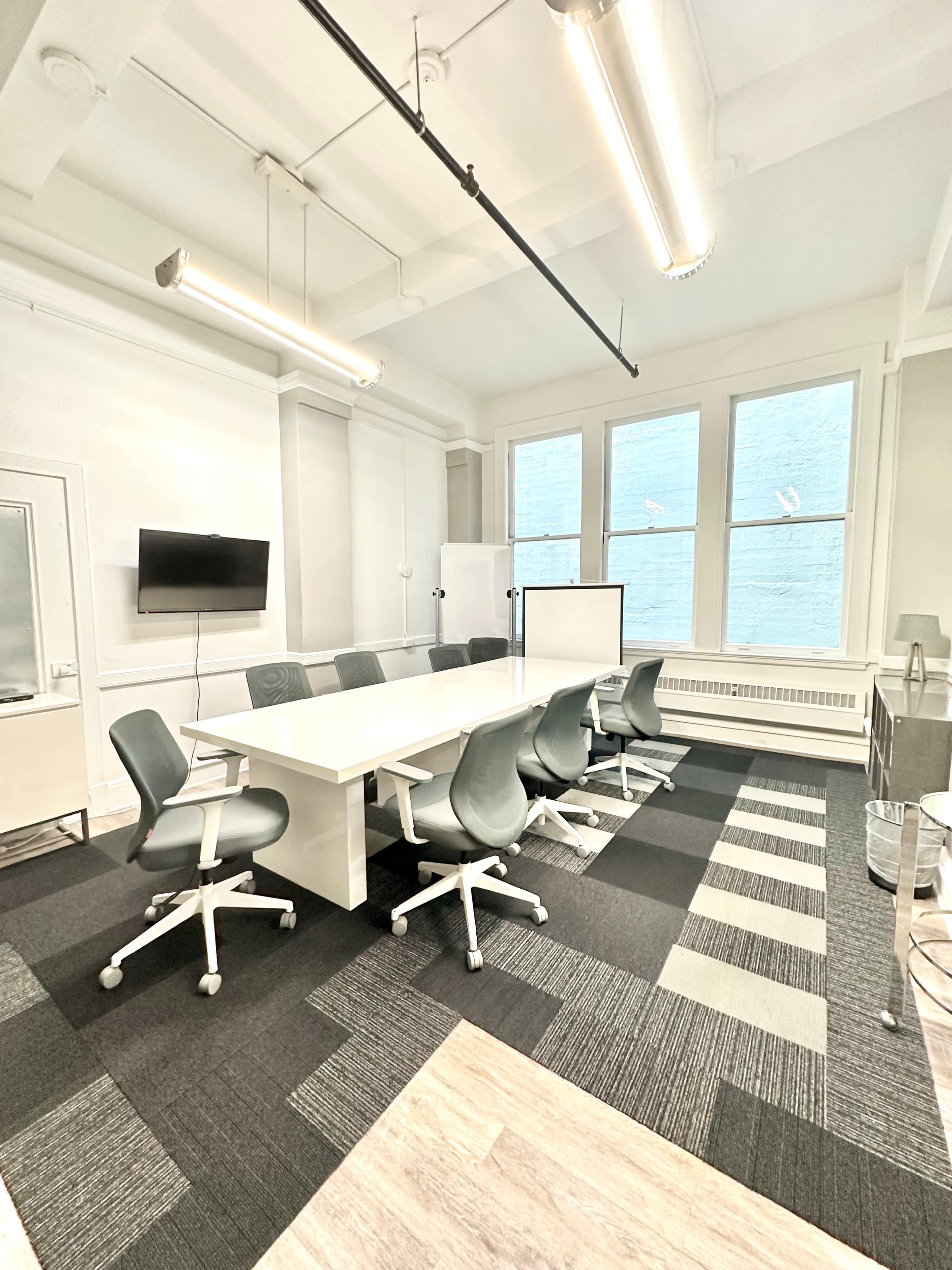 The image shows a modern conference room with a long white table, eight gray chairs, large windows, and a television mounted on the wall.