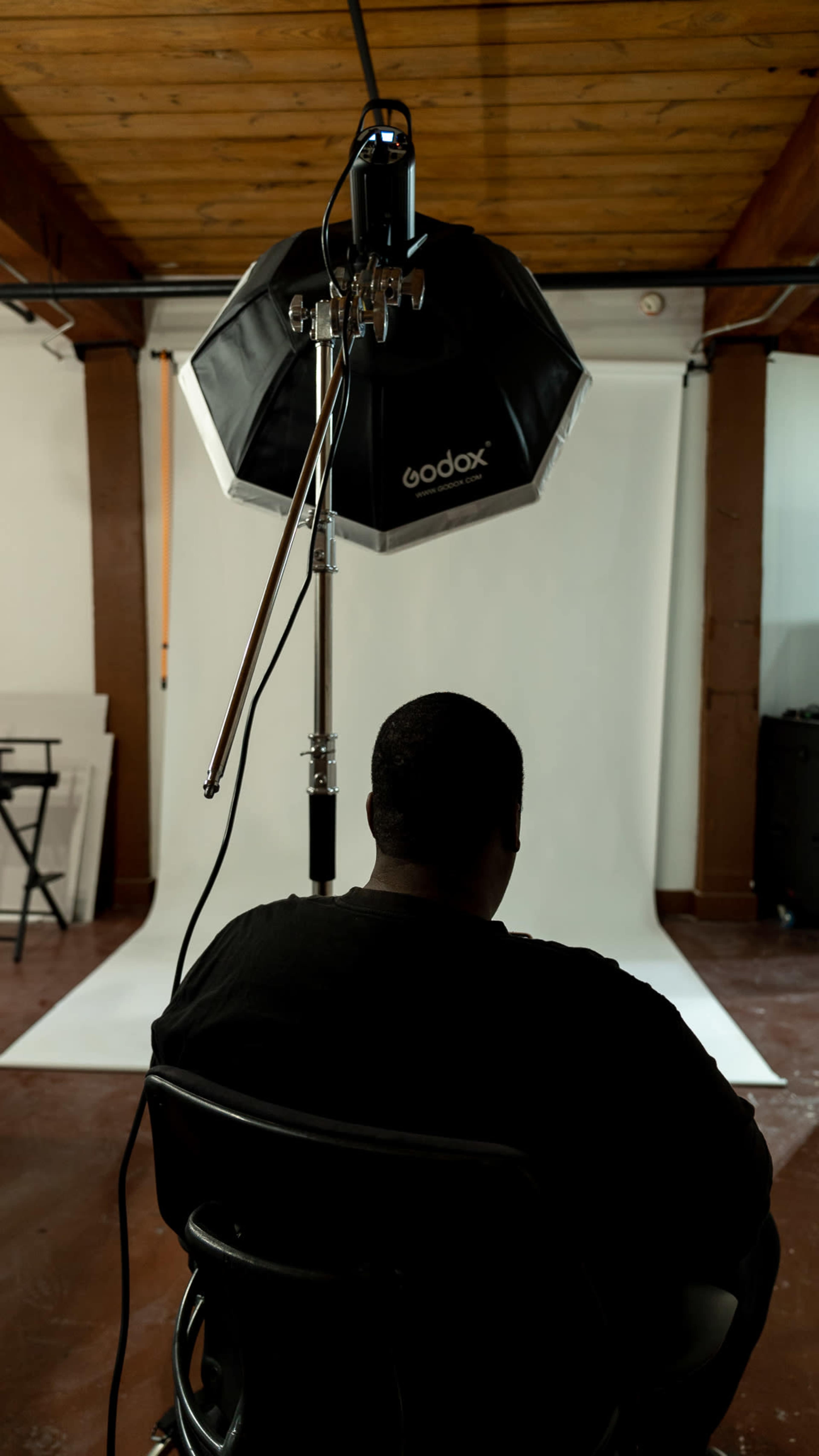 A person sits in a chair, facing away from the camera, under a large softbox light in a photo studio.