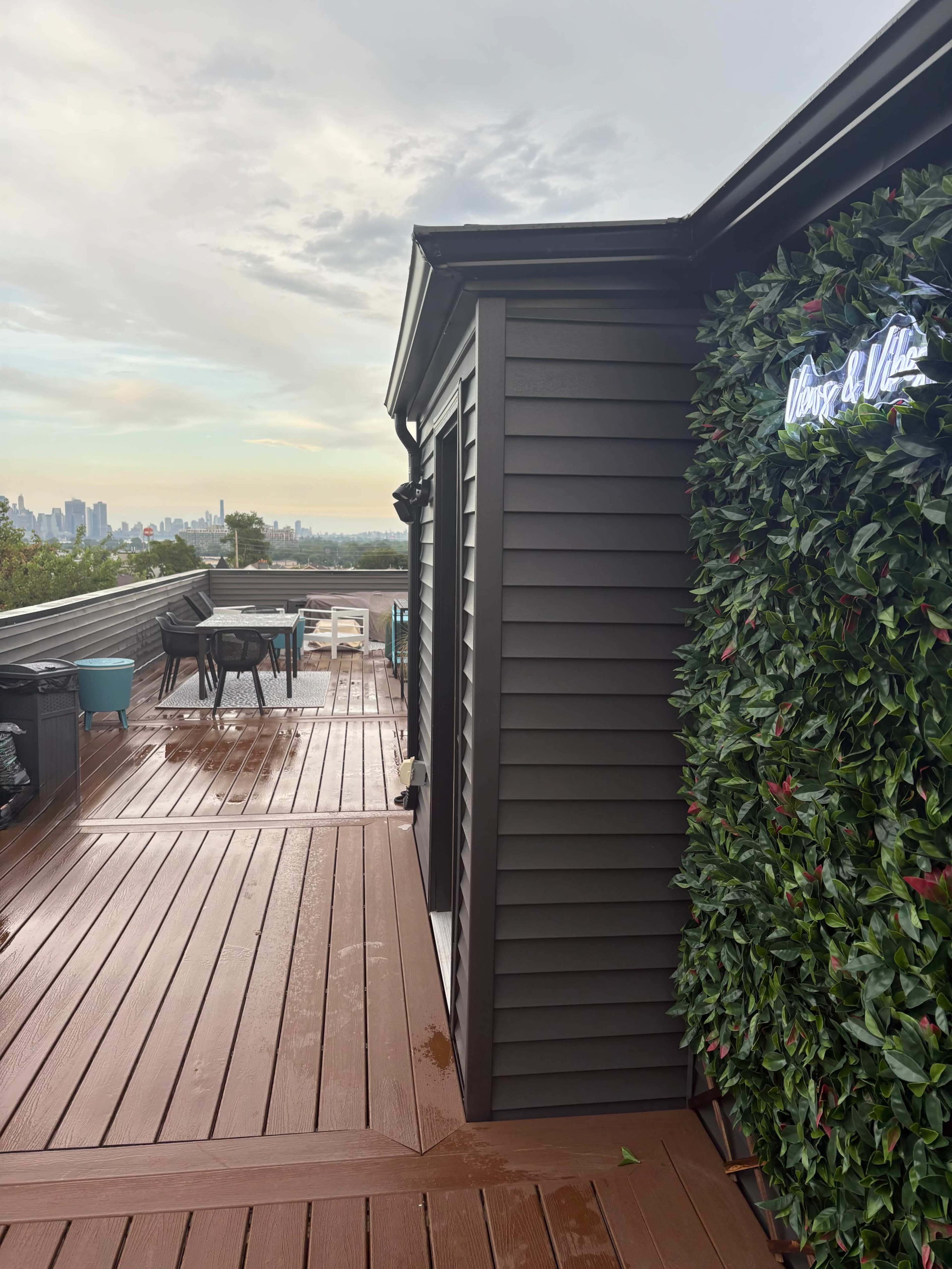 The image shows a rooftop deck featuring wooden flooring, a green wall of foliage, and a small table set with chairs, with a skyline view in the background.