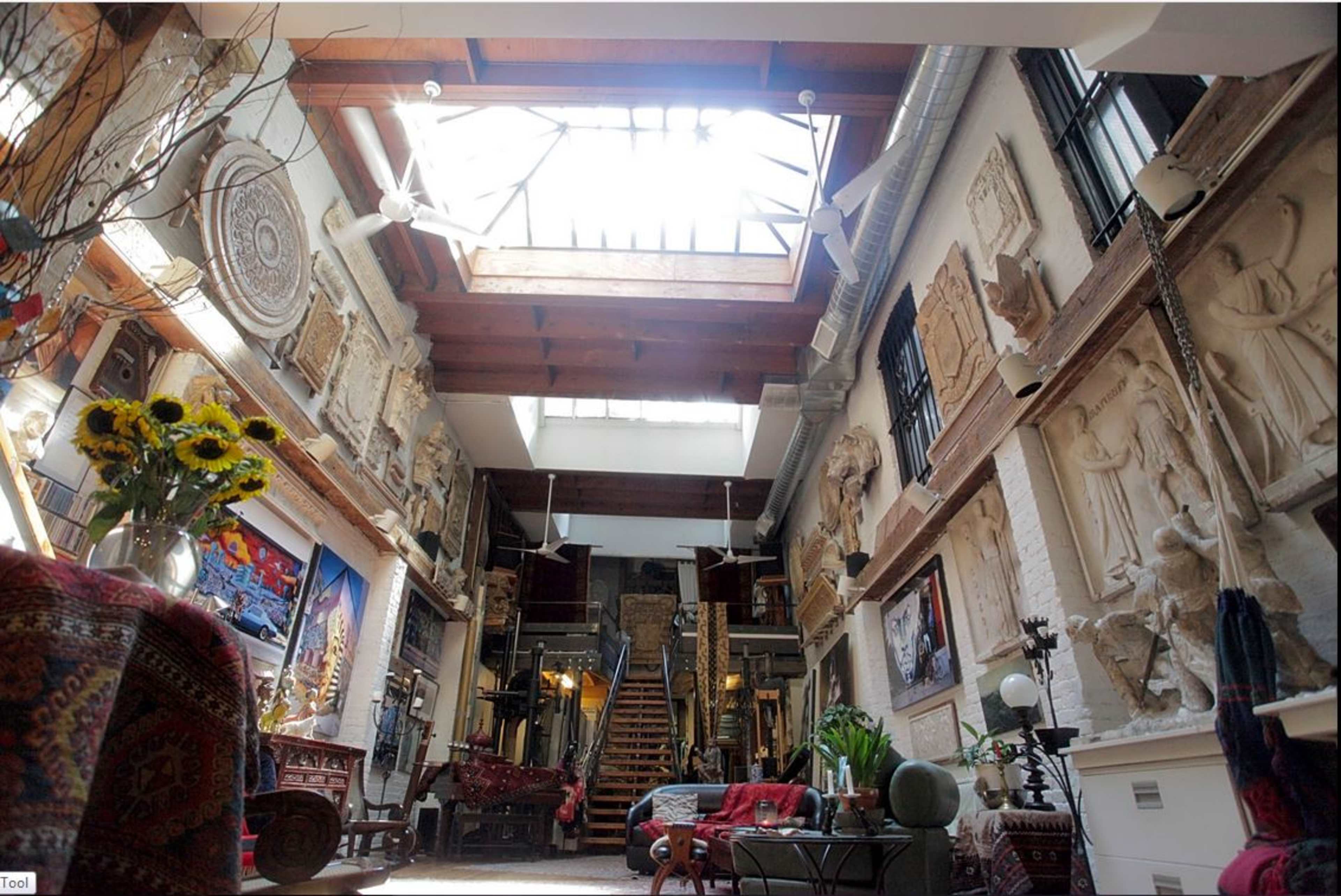 The image features a spacious interior with high ceilings, adorned with decorative wall art and sculptures, leading to a staircase at the back illuminated by natural light from a skylight.