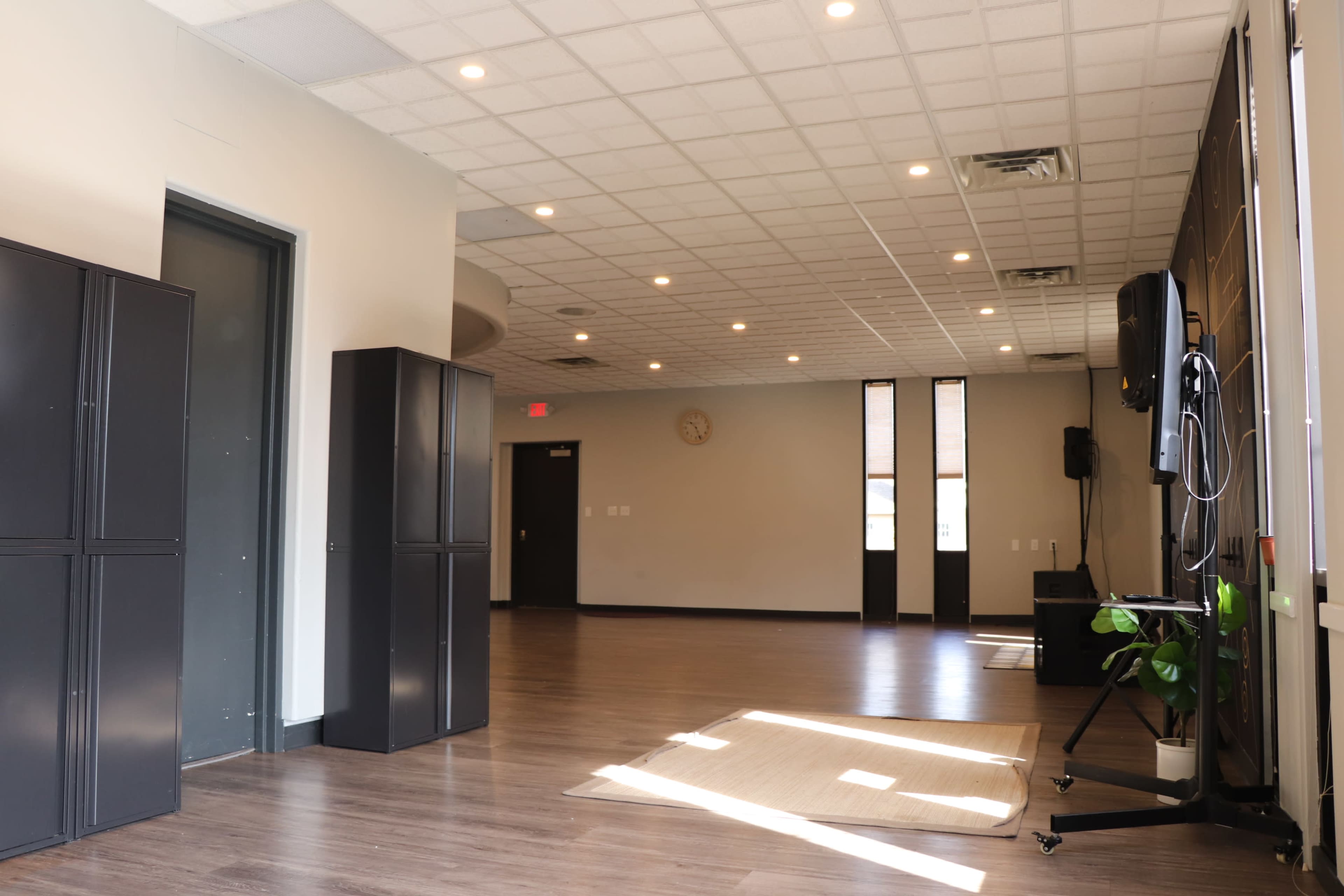 The image shows a spacious, well-lit room with gray storage lockers on one side and a television setup against the wall, featuring a rug on the floor.